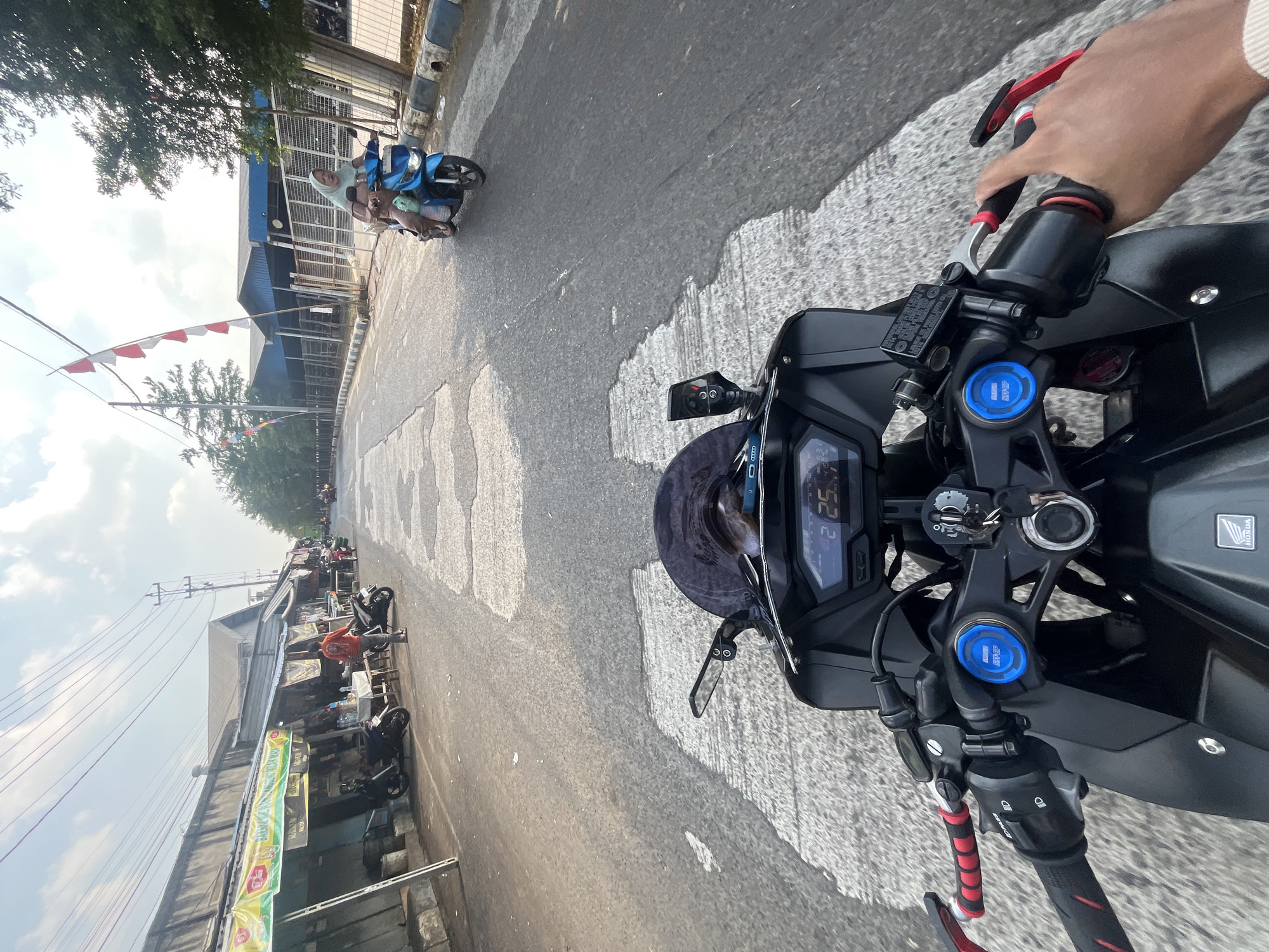 The image captures a first-person perspective from a motorcycle on a street in Pelang, Indonesia. The road is paved and shows signs of wear, with lighter colored patches indicating repairs or water damage. The left side of the street features a row of small buildings with awnings, some displaying signs and offering goods. Several motorcycles are parked along this side. On the right side of the road, trees line the pavement, and further back, industrial-looking buildings are visible. A blue scooter carrying two people, one in a headscarf, is traveling in the same direction as the viewer. Red and white Indonesian flags are strung across poles on the right side of the street, suggesting a national celebration or event. The viewer's hands are visible on the handlebars of the motorcycle, which has a digital display showing "2 25.4". The overall impression is a snapshot of daily life and local atmosphere in this Indonesian town.
