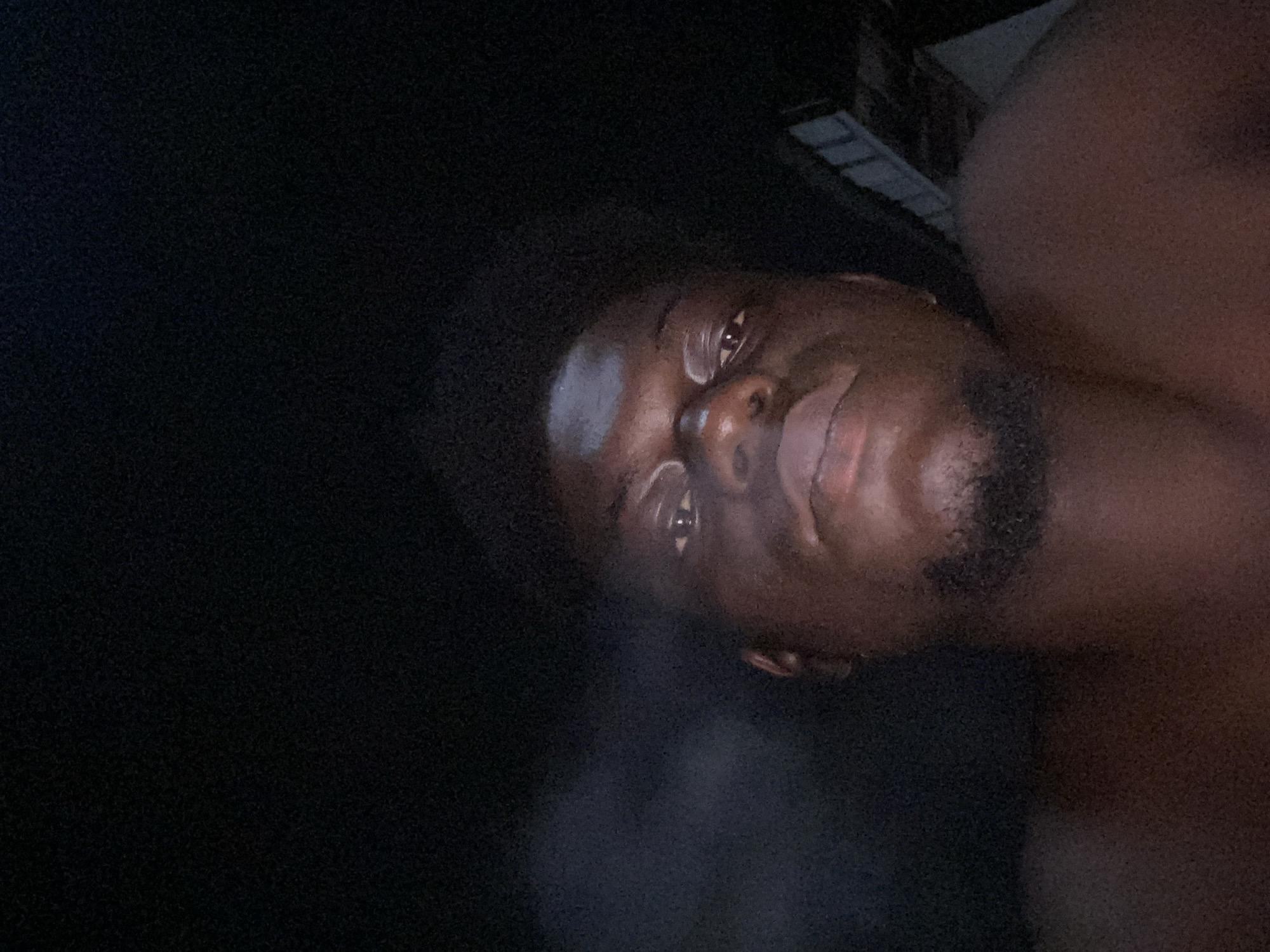A low-light photograph shows a dark-skinned person with a beard and short hair, possibly indoors based on a faint light source at the top right. The subject is facing the camera but the image is rotated counterclockwise, obscuring the background. The location cannot be determined.