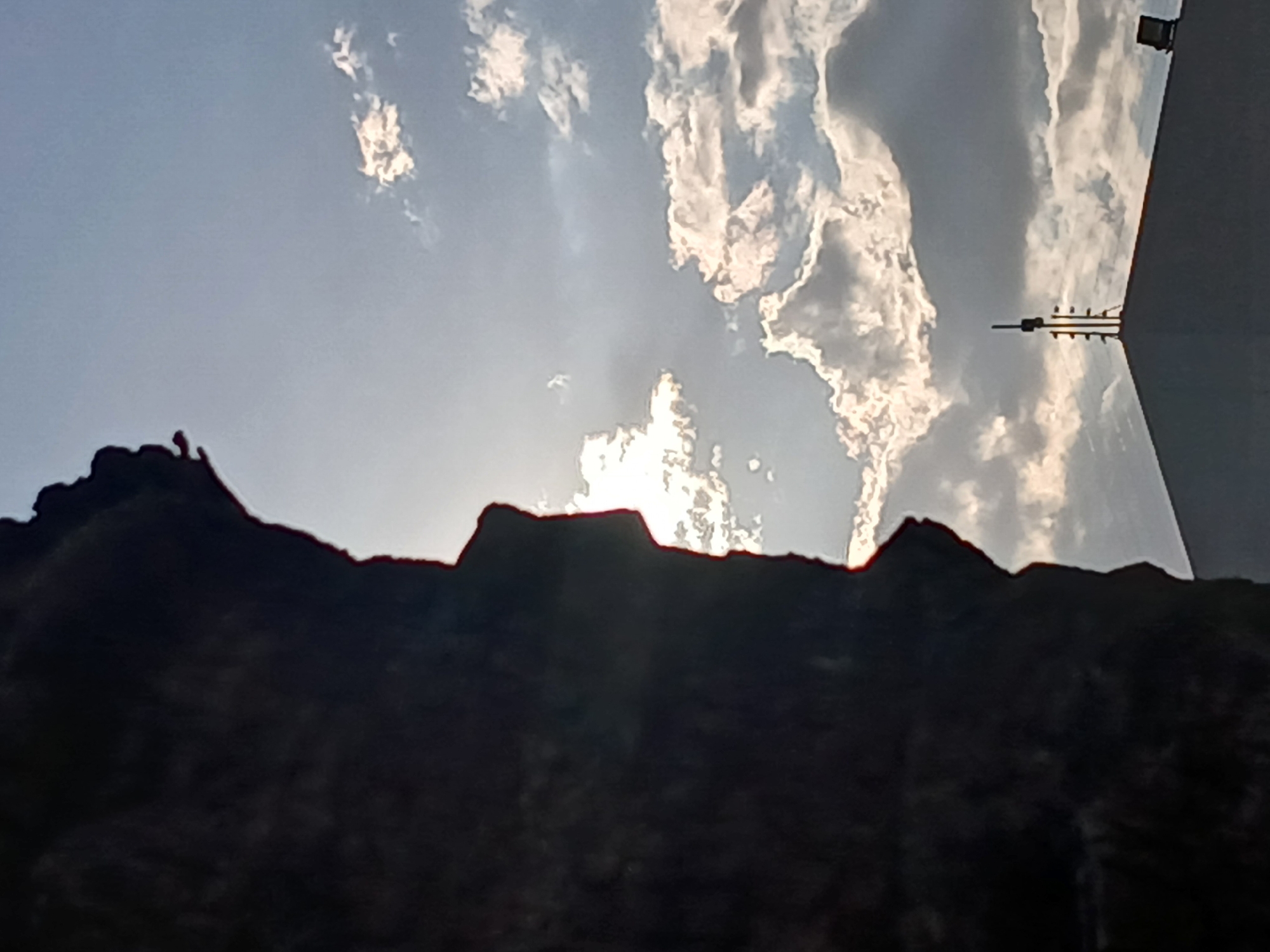 The image, when properly oriented, captures a dramatic landscape scene during either sunrise or sunset, characterized by strong backlighting and prominent silhouettes. The upper portion of the frame is dominated by a light blue to grey sky, filled with dynamic, textured cumulus clouds. These clouds are brilliantly illuminated from behind by the sun, which is mostly obscured by the cloud cover and the foreground elements, casting a radiant glow from the center-left.
Stretching across the lower-left is the dark, jagged silhouette of what appears to be a mountain range or rugged hills. The details of these natural formations are completely swallowed by shadow, appearing as a stark, dark mass against the bright sky.
On the right side, a sharp, dark silhouette of a building or a high wall/roof line is visible, suggesting an urban or developed area. Perched atop this structure is a distinct, dark antenna, possibly a television or communication aerial, with several horizontal crossbars, standing tall against the bright sky. A smaller, indistinct dark object, perhaps a utility unit, is also visible along the right edge of the building silhouette.
No specific activity or event is taking place; the image is a static portrayal of the environment. The time of day is clearly either early morning or late evening due to the powerful backlighting and deep shadows. The weather is partially cloudy, with the clouds contributing significantly to the atmospheric lighting. There are no visible people or text in the image. While no specific geographical markers are present, the scene depicts a blend of natural landscape and urban elements that could be found in a location such as Ciudad Guayana, Venezuela. The overall impression is one of striking natural beauty and dramatic light.