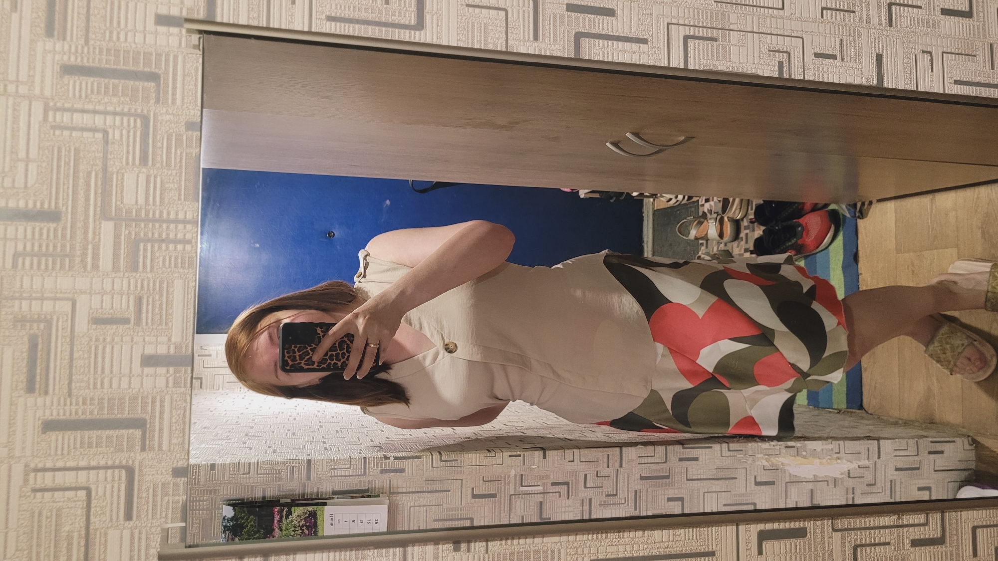 A woman stands in front of a mirror, taking a selfie. She is wearing a beige sleeveless top and a skirt with a geometric pattern in red, green, and white. Her hair is reddish-brown and cut short. She is holding a smartphone with a leopard-print case. The background appears to be an interior space, possibly a hallway or bedroom, with patterned wallpaper and a wooden closet door. A calendar is visible on the wall to the left of the mirror, and shoes are visible in the reflection of the mirror on the floor.