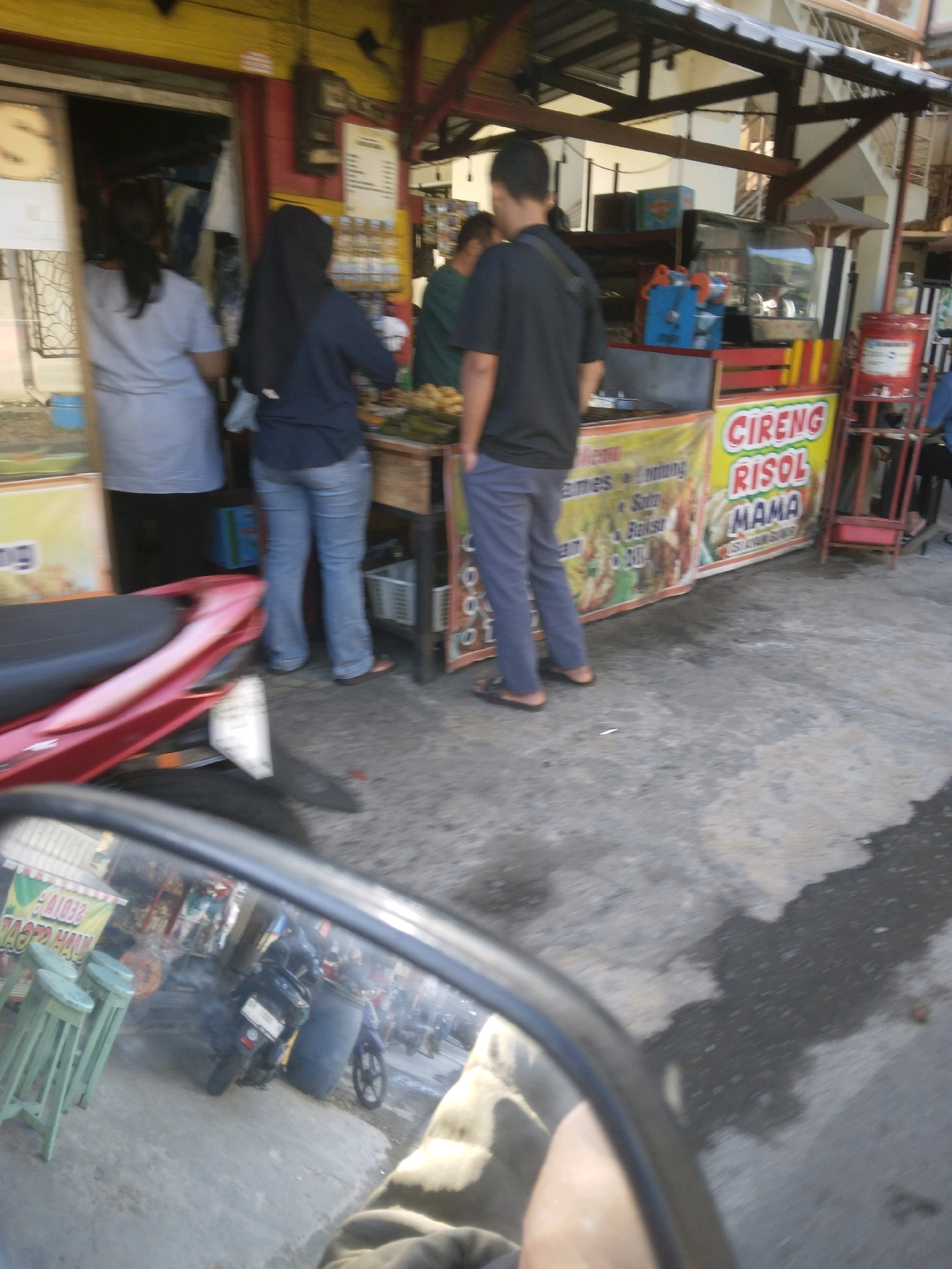 The image depicts an outdoor scene in Desa Pamijen, Kecamatan Sokaraja, Indonesia, likely a street or roadside food stall. In the foreground, a red motorcycle is partially visible, suggesting the photographer is on another vehicle or standing nearby. The main subjects are two women and a man. One woman, wearing a dark headscarf and dark top, stands in front of a food stall, facing away from the camera. Another woman, wearing a light-colored t-shirt, is also at the stall, her back to the camera. A man, wearing a dark t-shirt and blue pants, stands near the stall with his back to the camera. Behind them, the food stall displays food items and promotional banners, one of which reads "CIRENG RISOL MAMA." The environment appears to be a moderately busy street with concrete ground. The scene suggests a typical day at a local food vendor in Indonesia.