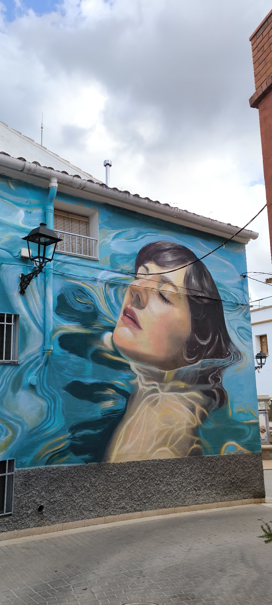 A mural depicts a woman's face partially submerged in water, rendered in a hyperrealistic style with soft lighting and vibrant blues. Her eyes are closed, and her lips are slightly parted. The water is characterized by swirling patterns and light reflections, creating a sense of depth and movement. The mural covers a significant portion of a building's exterior wall in Fanzara, Spain. The building itself is a two-story structure with a white roofline and a textured grey lower section. A black, ornate lamppost is mounted on the wall to the left of the mural, and a window with blinds is visible above it. Electrical wires run across the mural and the building. In the background, a red-brick chimney stack and a white building with a black lantern are visible. The ground in front of the building is paved with grey rectangular bricks. The sky above is partly cloudy.