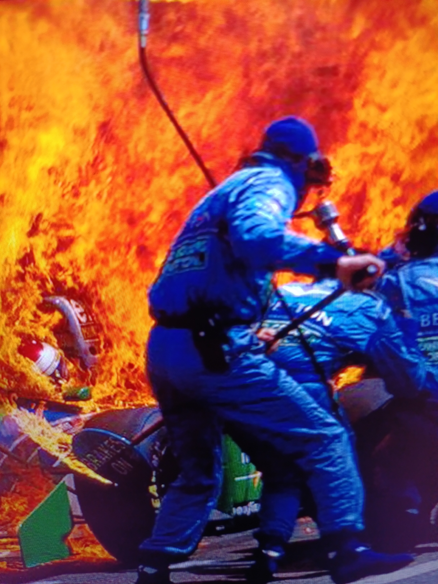 In a city scene, amidst intense flames and smoke, two individuals in blue racing suits are present. One individual, with their back to the camera and wearing a helmet, is holding a fire extinguisher hose, appearing to be actively fighting the fire. Another individual, also in a blue racing suit, is positioned to the right. In the foreground, a tire is visible with the words "BRAKES ON" printed on it. The environment suggests a chaotic and dangerous situation, likely a racing event incident.