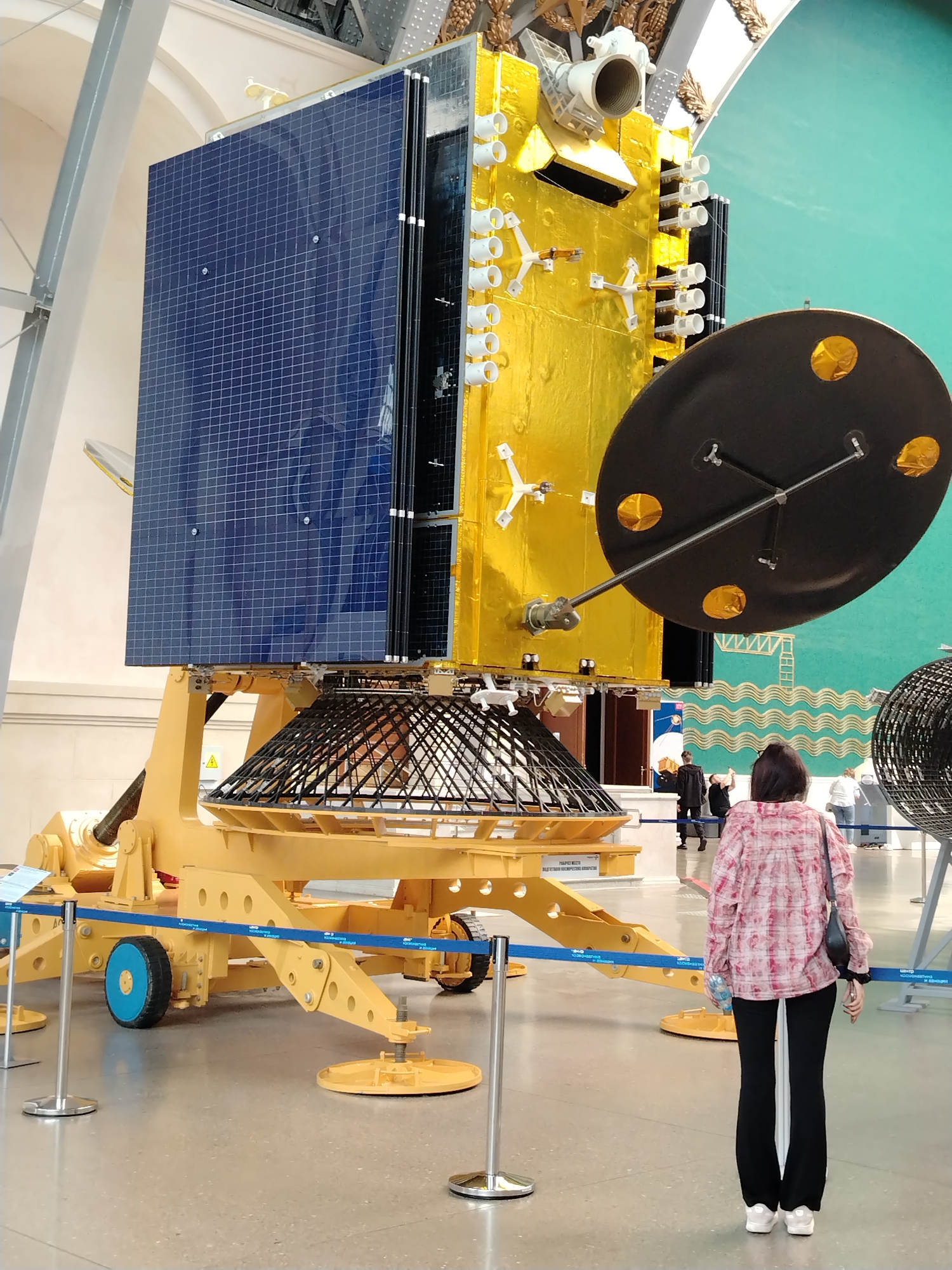 The image depicts a woman in a pink and white plaid shirt and black pants standing in front of a large satellite model, likely on display at a museum or exhibition. The satellite is predominantly gold and blue, with a large black dish antenna attached. It is mounted on a yellow, industrial-looking stand with wheels and stabilizers. The background suggests an indoor public space with a teal-colored wall featuring a wavy pattern. Other individuals are visible in the background, some interacting with exhibits. The overall impression is that of a public display of scientific or technological equipment. The presence of Cyrillic text on signage suggests a location in Russia, possibly Moscow.