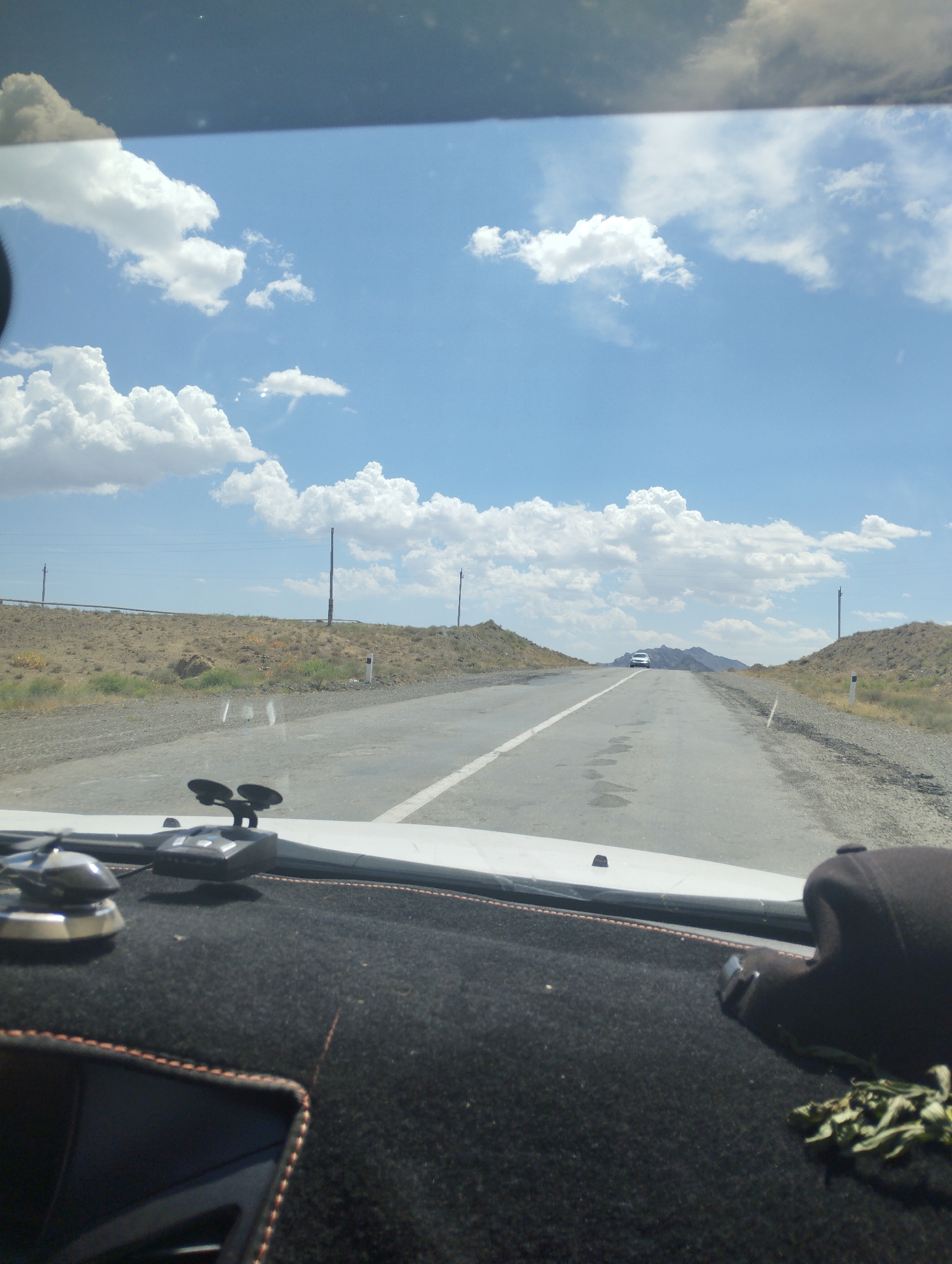 This is a forward-facing, first-person perspective image taken from inside a vehicle, likely on a paved road. The road stretches into the distance and appears to be a two-lane highway with a white dashed line dividing the lanes. On either side of the road are arid, dry hills with sparse vegetation. The sky is bright blue with numerous fluffy white clouds. In the distance, a white car is visible driving in the same direction. The foreground shows the dashboard of the vehicle, covered in a dark, textured material with orange stitching along the edges. Various accessories are mounted on the dashboard, including what appears to be a dashcam and a small metallic object. The overall environment suggests a rural or semi-arid location, possibly along a transportation route within Asmansay, Uzbekistan, given the dry landscape and typical roadside infrastructure like power poles.