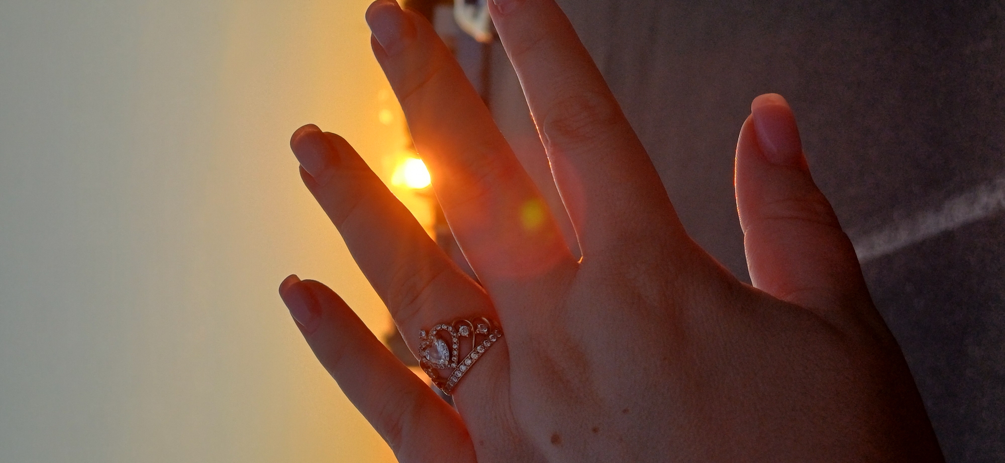 The image shows a close-up of a person's hand, with a ring on one finger, against a sunset sky. The ring is a rose gold crown-shaped ring with a central clear stone and smaller stones along the band. The background is a gradient of orange and yellow from the setting sun, with a hazy blur of what appears to be buildings or trees in the distance. The lighting suggests it is evening. The environment appears to be outdoors, possibly a street or parking lot, with a dark, asphalt-like surface visible in the lower part of the frame. There is no discernible information to suggest the location is the city of Kyiv, Ukraine.