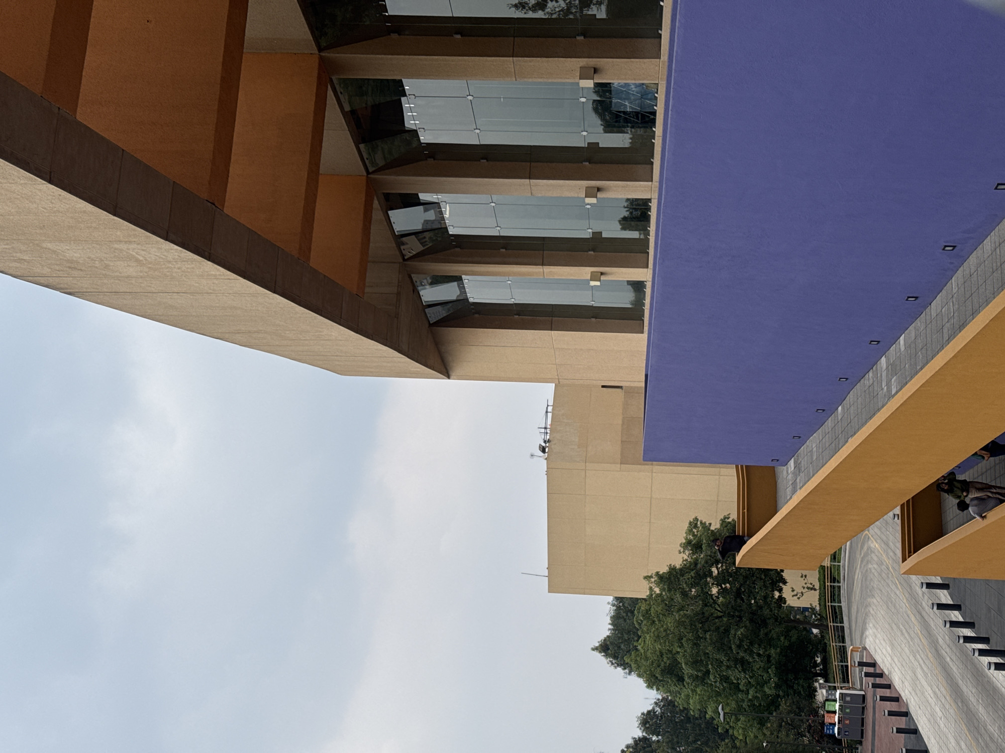 The image shows a section of the Universidad Nacional Autónoma de México (UNAM) campus in Mexico City. The architecture is modern, with concrete structures, glass windows, and bold colors like orange, purple and brown. The sky is visible in the background. Trees can be seen nearby, adding a natural element to the urban environment. The image appears to be taken from a low angle, emphasizing the scale and design of the buildings.