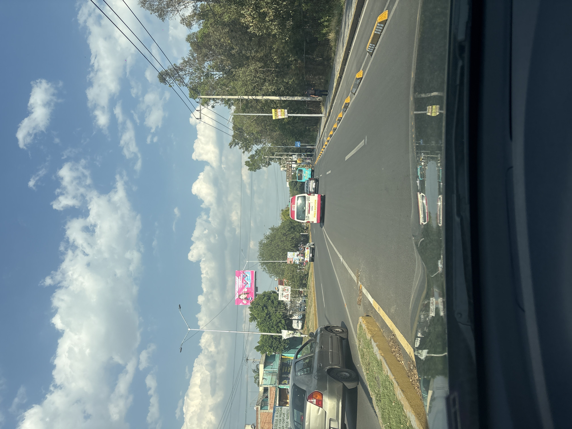 Captured from a vehicle's perspective, the image reveals a street scene in Puebla, Mexico. A multi-lane road stretches into the distance, bordered by trees and utility poles. Several vehicles, including a bus, a compact car, and a delivery truck, are visible on the road. Sidewalks line the road with green grass. Billboards and commercial signs mark the side of the road. The sky above is partly cloudy, providing diffused light.