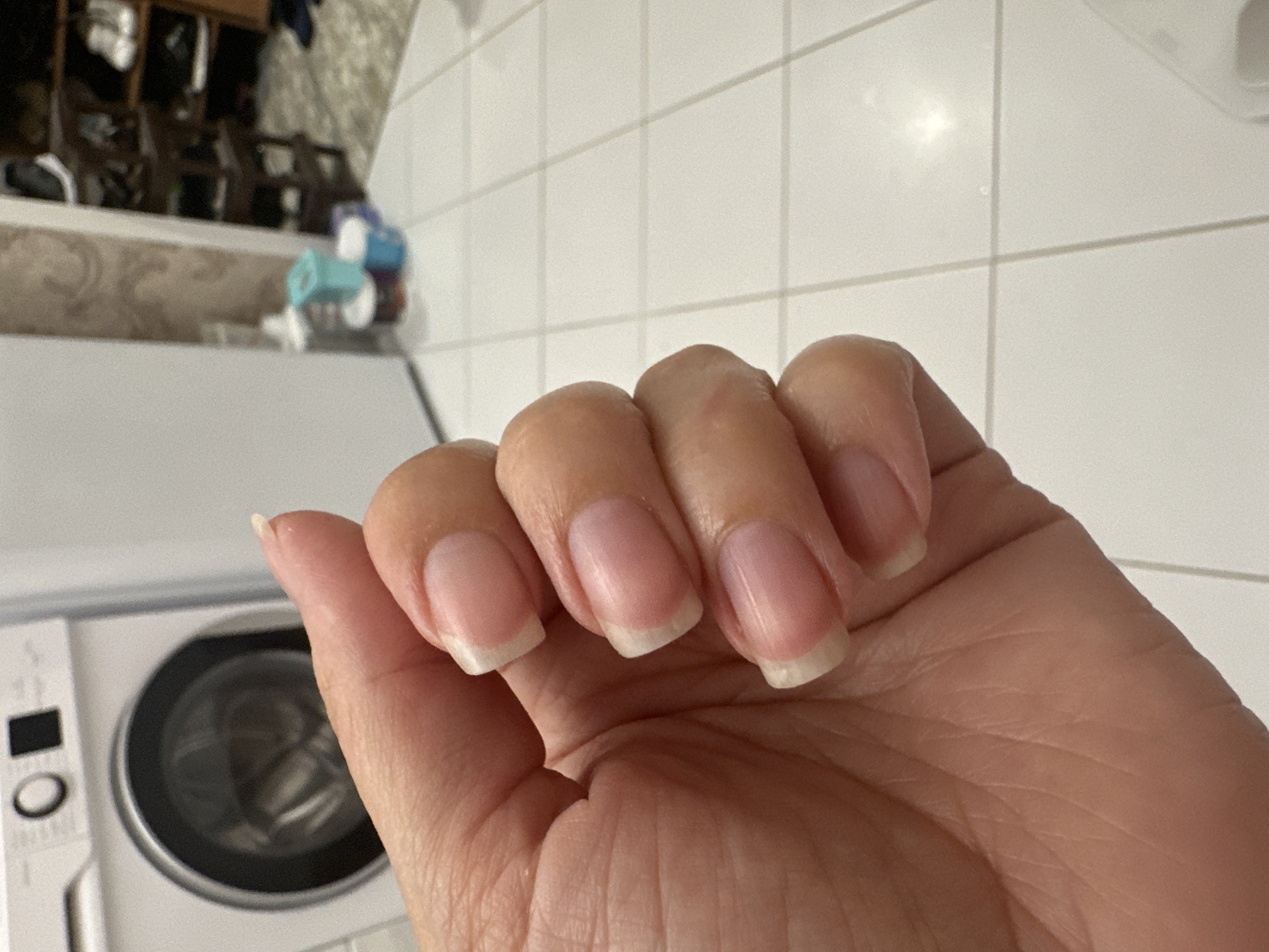 A close-up, eye-level, over-the-shoulder shot shows a person's left hand with natural, short, manicured fingernails. The hand is held up against a tiled floor and a blurred background. The fingernails are clean and appear healthy, with a light pink base and white tips, characteristic of a French manicure. The skin on the hand is smooth and unblemished.
In the background, a white washing machine is visible on the left, with its circular door slightly ajar. To the right of the washing machine, a white wall transitions into a patterned wallpapered area, with shelves of various items, possibly in a storage or laundry room. A few colorful containers are clustered on the floor near the wall. The floor is covered with light-colored square tiles, arranged in a grid pattern. The lighting is neutral, creating soft shadows. There is no indication of the specific location within Verkhniy Yurt, Russia.