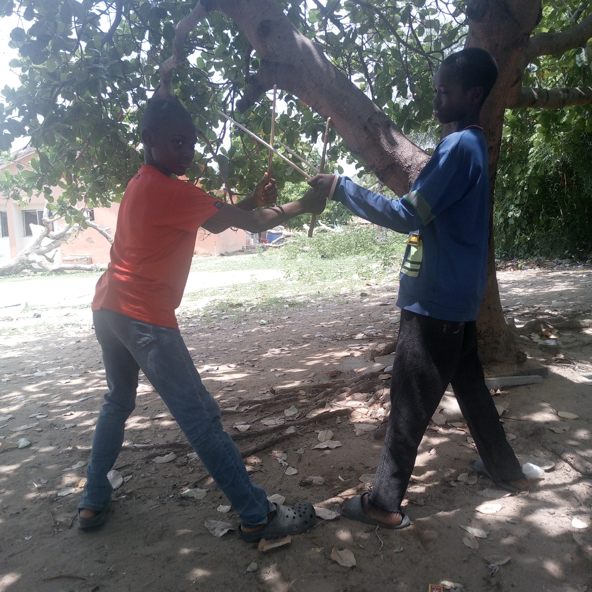 In Adankolo, Nigeria, two dark-skinned children stand beneath a tree, operating a homemade device constructed of sticks and string. The child on the left wears an orange t-shirt and blue jeans, while the child on the right is dressed in a blue long-sleeved shirt and black pants. Both are wearing sandals. They face each other, holding the ends of the device, which consists of two upright sticks connected by taut strings. The ground is dirt and scattered with leaves, and a building is visible in the background. The scene is dappled with shadows cast by the tree's foliage.