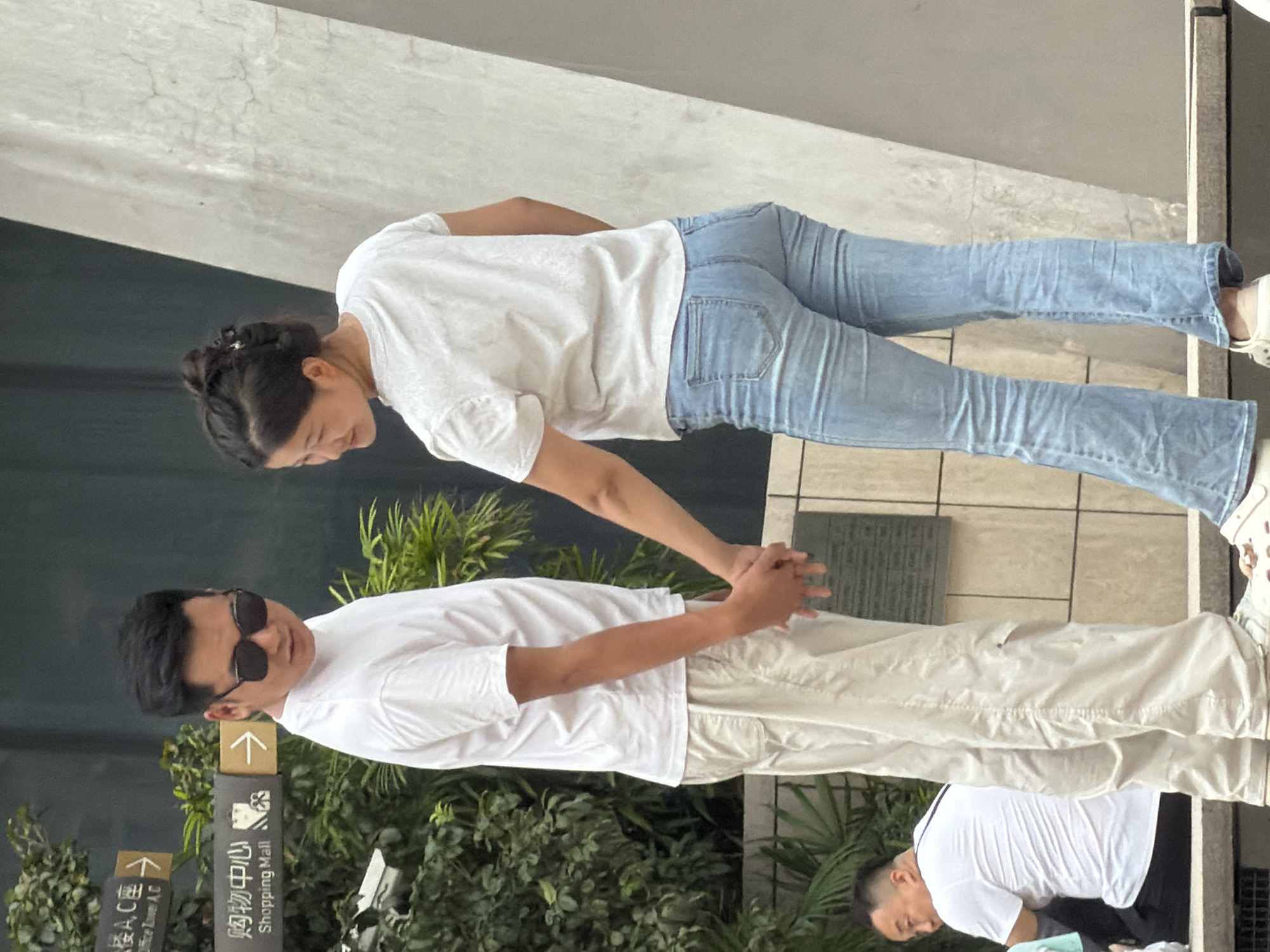 In Yuzhong District, China, a man and a woman are holding hands and facing each other in an outdoor urban environment. The man, wearing sunglasses, a white t-shirt, and light-colored pants, is on the left. The woman, with her hair in a bun, is wearing a white t-shirt and light blue flared jeans. Behind them, there are trees and signage indicating "Shopping Mall." To the left of the couple, another man in a white t-shirt is seated. The background features a textured concrete wall.
