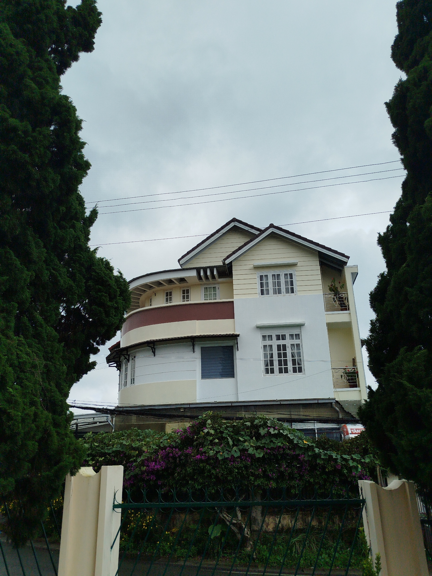 The image depicts a multi-story building in Xuân Hương - Đà Lạt, Vietnam, characterized by its curved facade and white and cream colored exterior. The building features multiple windows, some with a grid pattern, and balconies with railings. It is surrounded by lush green trees on either side, framing the structure. In the foreground, a beige pillar and a green metal fence are visible, with dense vegetation and purple flowers growing behind the fence. The sky is overcast and gray. The scene appears to be a residential or commercial property situated within a natural, possibly mountainous, landscape.