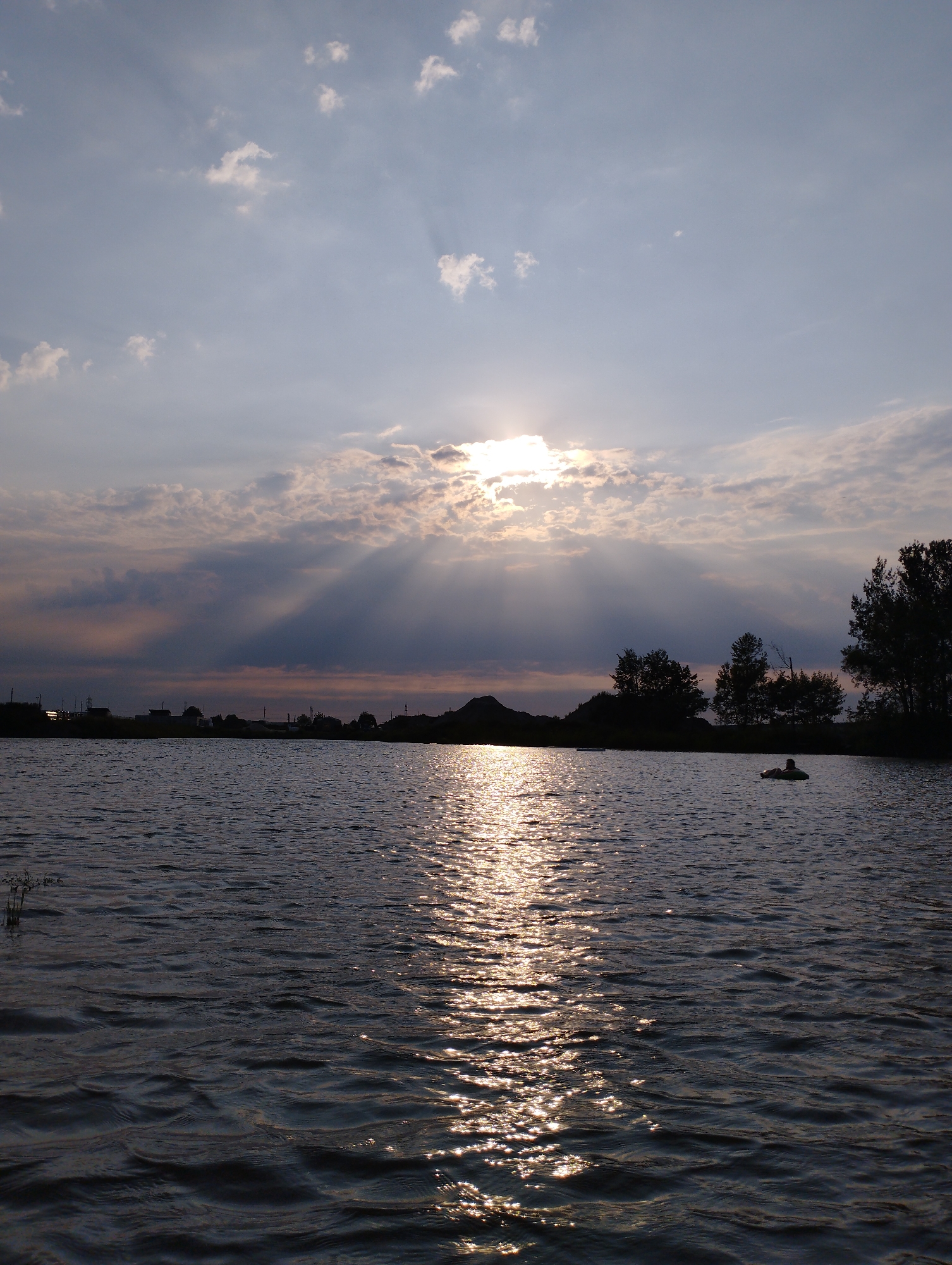 The image depicts a sunset over a body of water, likely a lake or river, with a silhouette of trees and buildings along the horizon. The sun is partially obscured by clouds, casting dramatic sunbeams through the sky. The water surface reflects the sunlight, creating a shimmering path. In the water, a person is visible in an inflatable tube, appearing to be relaxing or swimming. The scene suggests a tranquil, natural setting within or near a populated area. The location is confirmed as Michałowice-Wieś, Poland.