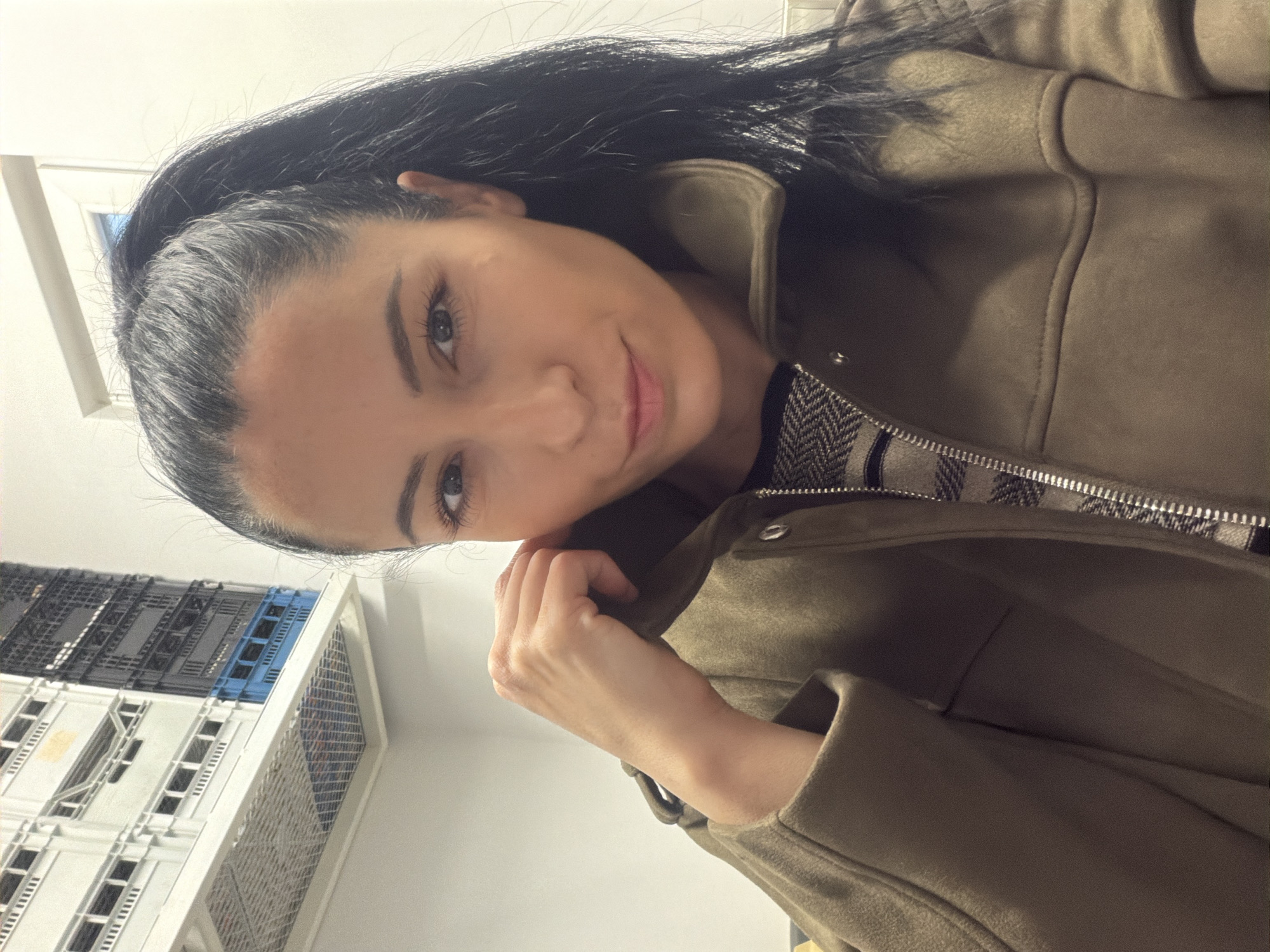 A woman with long black hair and fair skin, wearing a brown jacket with a patterned lining, poses for a selfie. The background includes a white wall and a structure with rows of compartments, some of which are blue. The location is unspecified and cannot be determined from the provided image.