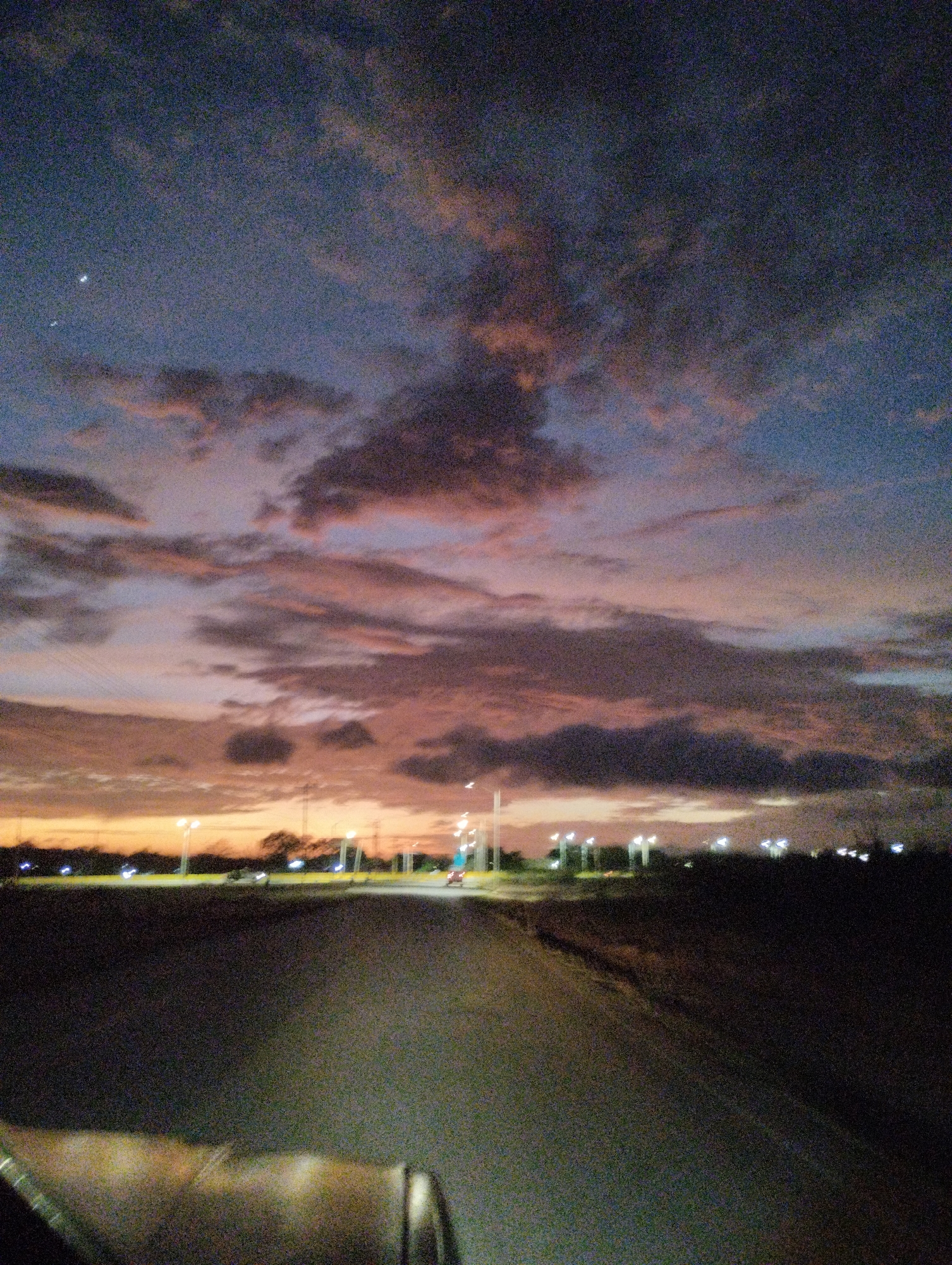 The image depicts a twilight scene with a dramatic sky filled with clouds in various shades of purple, pink, and gray. Two celestial bodies, possibly the moon and a planet, are visible in the upper left quadrant of the sky. A road stretches into the distance, bordered by dark land on either side, with a line of streetlights illuminating the horizon. A few distant trees and silhouetted structures can be seen along the horizon. The foreground shows a portion of what appears to be a vehicle's interior, suggesting the photo was taken from a moving car. The overall atmosphere is serene and atmospheric, characteristic of a rural or semi-rural setting in the vicinity of La Guardia, Venezuela, during dusk.