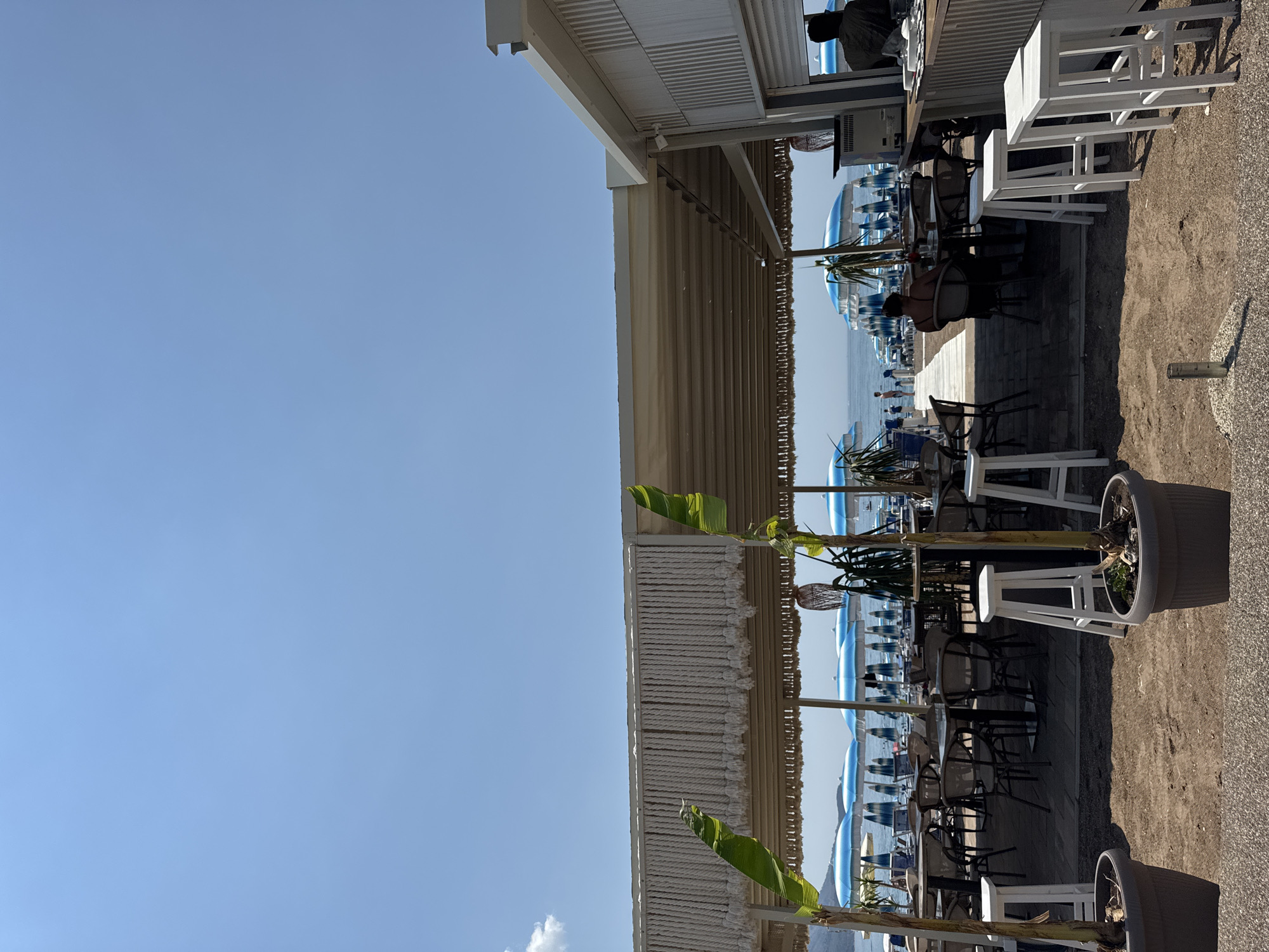The image depicts an outdoor seating area of a beachside establishment, likely a cafe or restaurant, located in Boreti, Montenegro. The scene is set on a sunny day, as indicated by the bright blue sky with a few wispy clouds and the strong shadows cast by the furniture. The time of day appears to be late morning or early afternoon.
In the foreground, there is a gravel and sandy area with several white bar stools and tables. Beyond this, an open-air covered area with a white slatted roof and woven decorative elements provides shade for a collection of tables and chairs. Some of these tables are occupied by patrons, including a person sitting at a table and looking out towards the sea, and another person at the counter.
The background reveals a beach with numerous blue and white beach umbrellas and lounge chairs, suggesting it is a popular spot. The calm sea stretches towards the horizon under a clear sky. Lush green tropical-looking plants, possibly banana trees with large leaves, are interspersed throughout the seating area, adding a touch of tropical ambiance. The overall mood is relaxed and leisurely, typical of a beach vacation setting. There is no visible text in the image.