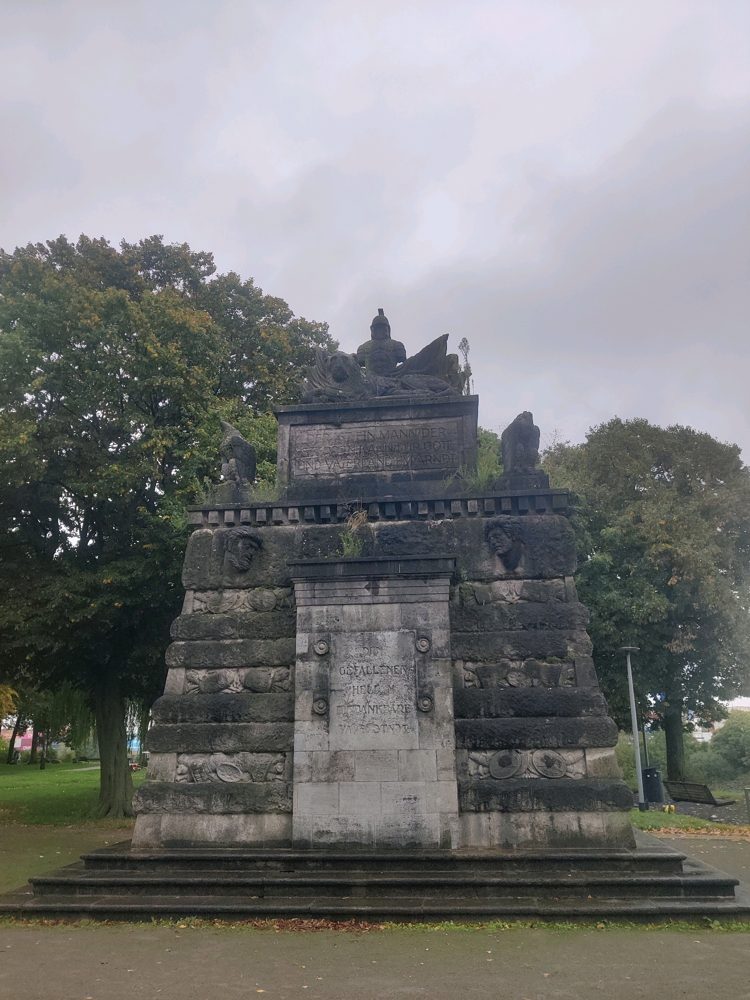This image shows the Bismarck Monument in Emmerich am Rhein, Germany. The monument is made of stone and is topped with a statue of Bismarck. The monument is located in a park, with trees in the background. The sky is overcast.