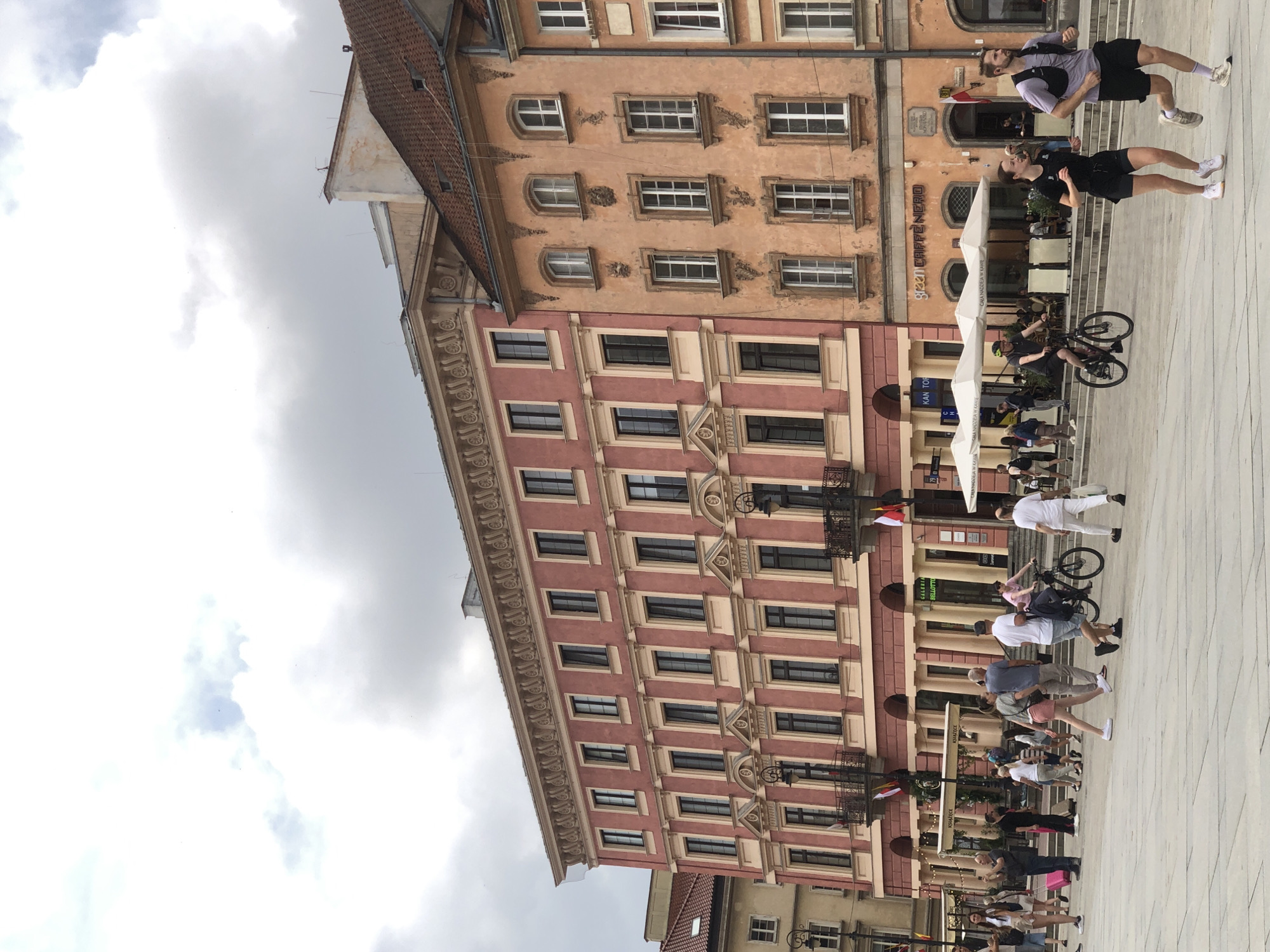 This is an outdoor, eye-level, full shot of a city square in Warsaw, Poland, on a cloudy day. In the background, there are two multi-story buildings. The building on the left is a salmon-pink color with white trim around the windows and cornices. It has numerous windows and ornate architectural details. The building on the right is a pale orange color with white trim and similar window and cornice detailing. It also features arched windows on its upper floors. Below the buildings, there are several cafes with outdoor seating under umbrellas.
In the foreground, there is a paved plaza with people engaged in various activities. A couple is running towards the right side of the frame; the man is wearing a purple t-shirt and black shorts, and the woman is wearing a black athletic top and shorts. Behind them, a man is riding a bicycle. Other individuals are walking or standing around the plaza. The ground is light-colored paving stones. The overall impression is a bustling urban environment.