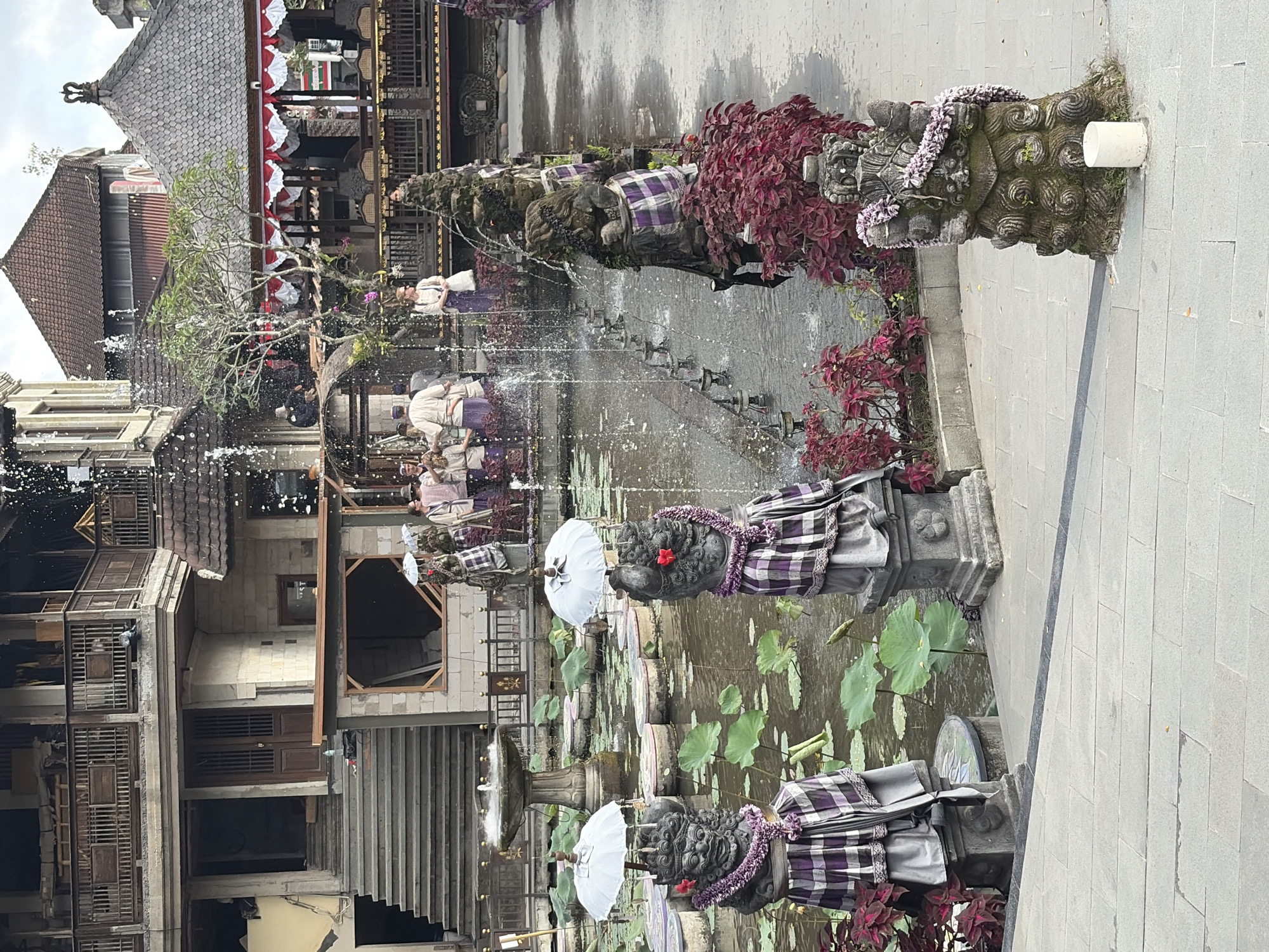 This is a medium shot, eye-level, outdoor photograph of a water feature in the city of Ubud District, Indonesia. In the foreground, two stone statues are adorned with purple and white checkered fabric and floral garlands. These statues flank a stone path leading to a pond with lily pads and a fountain in the background. A group of people in traditional attire are visible near the fountain, some holding white umbrellas. The architecture in the background includes multi-story buildings with balconies and tiled roofs, typical of Balinese design. The overall environment appears to be a cultural or historical site, possibly a temple or palace complex. The lighting suggests it is daytime, with overcast or diffused light.