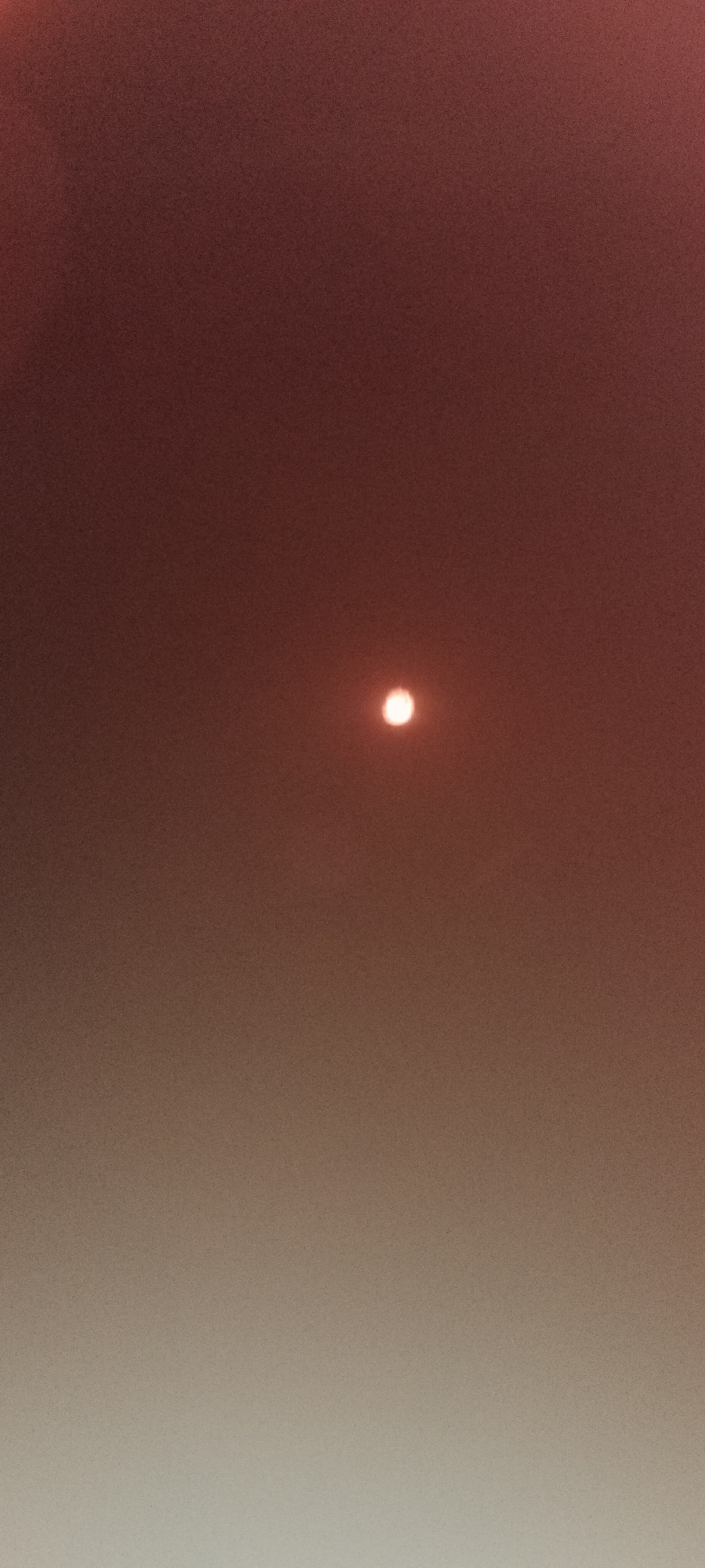 The image shows a hazy reddish-orange sky with a bright, out-of-focus circle in the center, likely representing the sun obscured by atmospheric conditions. The sky transitions from a darker reddish hue at the top to a lighter, more muted tone towards the bottom, suggesting a gradient of light or haze density. There are no discernible subjects like people or objects, and the scene is abstract, dominated by the sky.
The overall impression is that of a sunrise or sunset, possibly impacted by smoke, dust, or pollution, creating a diffused and colored sky. The out-of-focus sun adds to the ethereal quality of the image. No text is visible. The setting appears to be outdoors, given the sky, and the time of day is likely dawn or dusk due to the quality of light and color. The location context of Adai, Spain, could imply a Mediterranean climate, but the image itself does not provide specific visual cues to confirm this. The muted colors and haze might evoke a sense of calm, mystery, or even a slight unease depending on the viewer's interpretation of the atmospheric conditions.