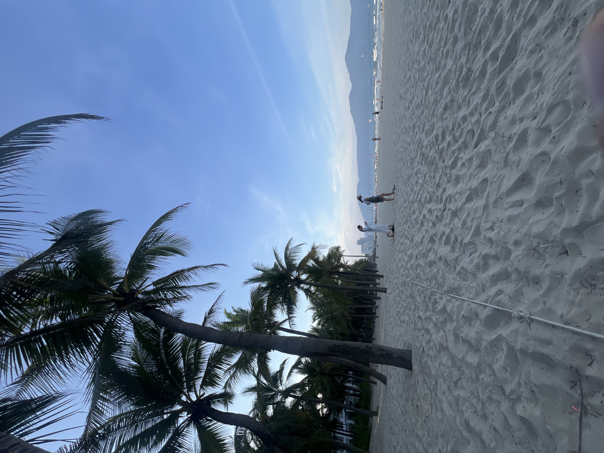 A beach scene in Đà Nẵng, Vietnam, features a wide expanse of light-colored sand under a clear blue sky with wispy clouds. Several palm trees with dark green fronds are visible in the foreground, their trunks leaning towards the left of the frame. In the mid-ground, a line of palm trees creates a natural border, with what appears to be buildings or resorts visible behind them. Further in the distance, mountains form a hazy backdrop against the horizon.
Two people are present on the beach. One person, wearing a hat and shorts, is captured mid-stride on the sand. Another person, dressed in white, stands near the shore. The presence of a rope stretching across the sand suggests some form of boundary or activity. The lighting indicates it is late afternoon or early evening, with the sun casting long shadows. The overall atmosphere is peaceful and relaxed, typical of a beach setting. There is no visible text in the image.