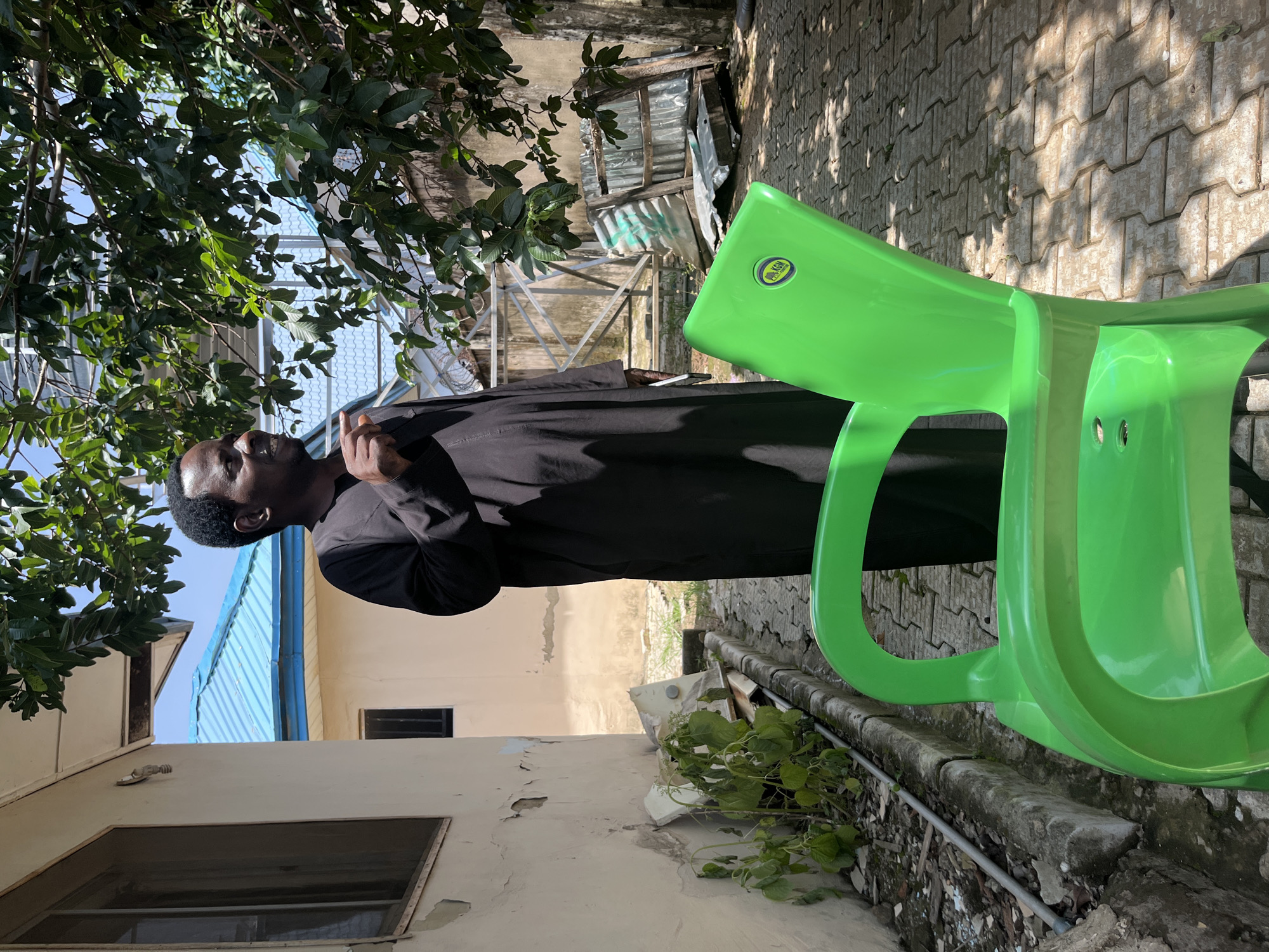 A man in a black traditional outfit stands outdoors in a courtyard. He is looking to the right and gesturing with his right hand. A bright green plastic chair is in the foreground, obscuring the lower part of his body. The courtyard is paved with interlocking bricks and has a drain running along the side, next to a wall with peeling paint. There are some plants growing by the wall and some debris in the background, including corrugated metal sheets and wooden planks. Behind the man, a building with a blue metal roof is visible. The scene appears to be in a residential area of Abuja, Nigeria.
