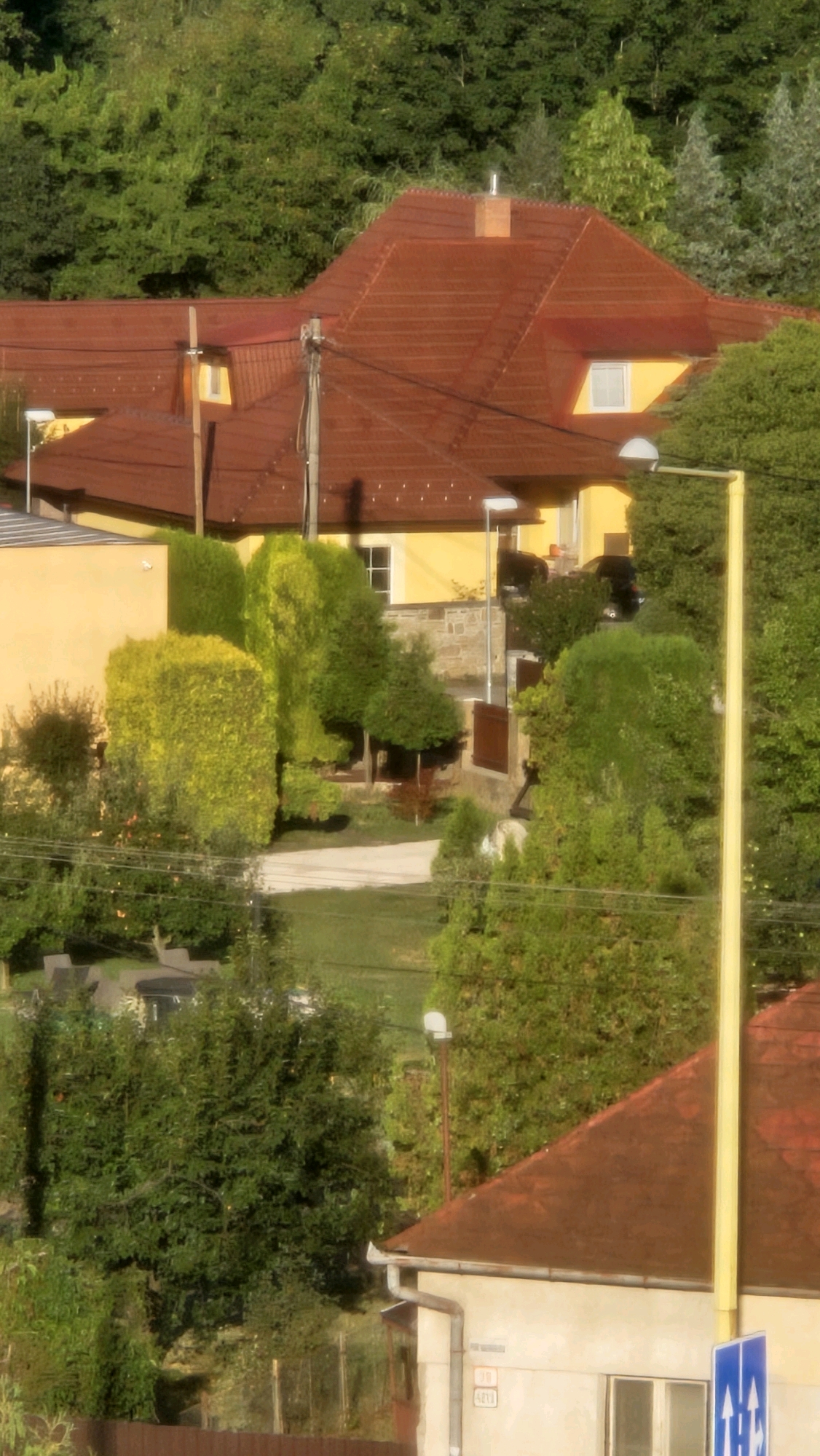 The image captures a residential area in the city of Prešov, Slovakia, likely a suburban or semi-rural setting, characterized by houses with prominent reddish-brown tiled roofs and lush green trees surrounding them. The foreground features a portion of a light-colored house with a prominent gutter system and a blue street sign with white arrows indicating directions. Adjacent to this house, a tall, bright yellow pole stands vertically. Behind this structure, a dense line of trees forms a backdrop.
Further into the scene, more houses with similar reddish-brown roofs are visible, interspersed with abundant vegetation. One house is painted yellow and features a red-tiled roof. Several streetlights are present throughout the scene, some mounted on poles and others appearing to be attached to the houses. The overall impression is that of a tranquil, green residential neighborhood on a sunny day, with evidence of modern infrastructure like streetlights and signage. The presence of numerous trees suggests a location with significant green space.