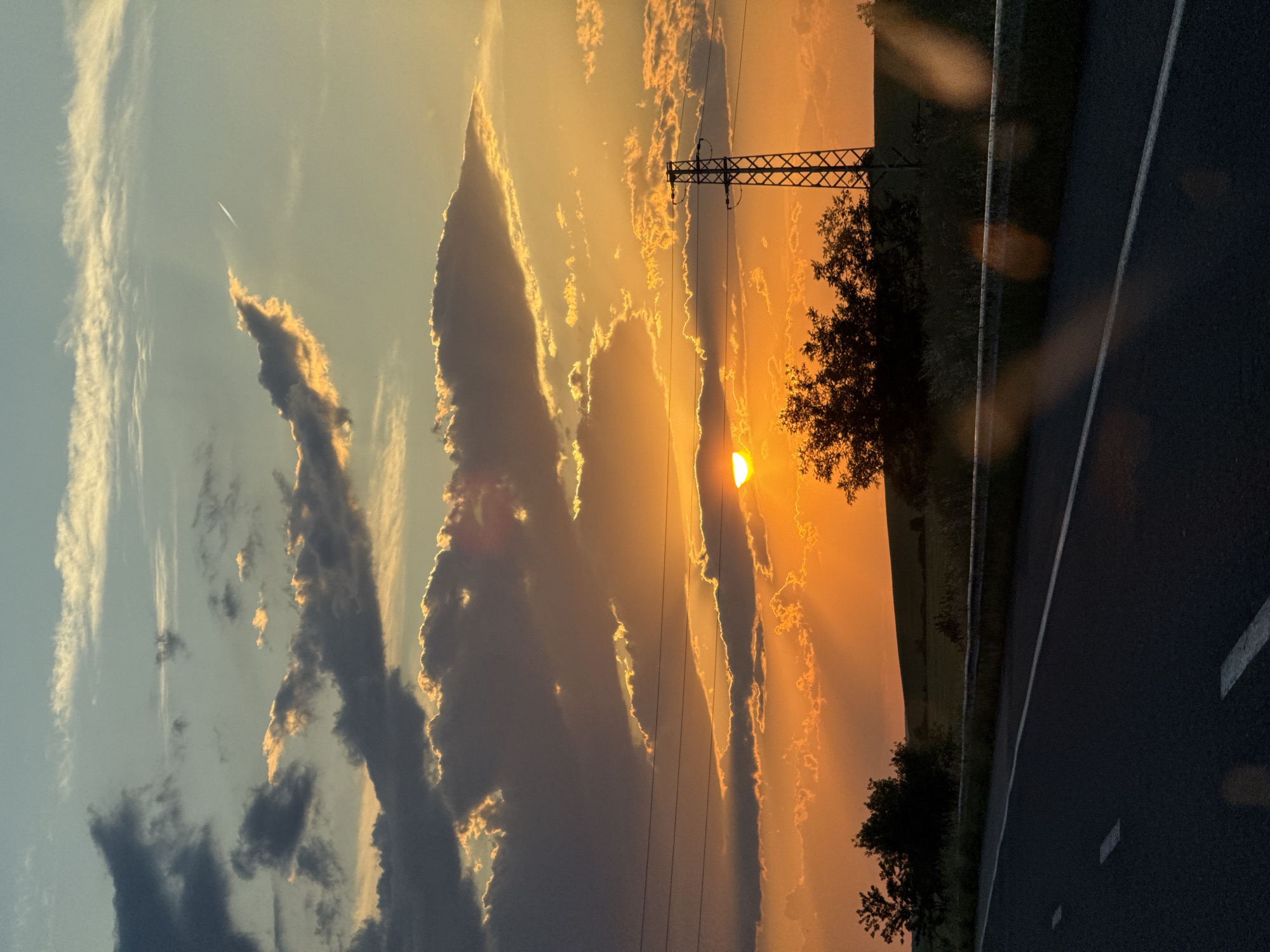 A photograph depicts a vibrant sunset, partially obscured by dark clouds, with beams of light breaking through. A utility pole with wires is visible near the center of the frame. Trees are silhouetted against the bright sky. A portion of an asphalt road with white lines runs along the bottom right of the image. There is no evidence to confirm this picture was taken in the city of Zetiovo, Bulgaria. No people are visible in the frame.