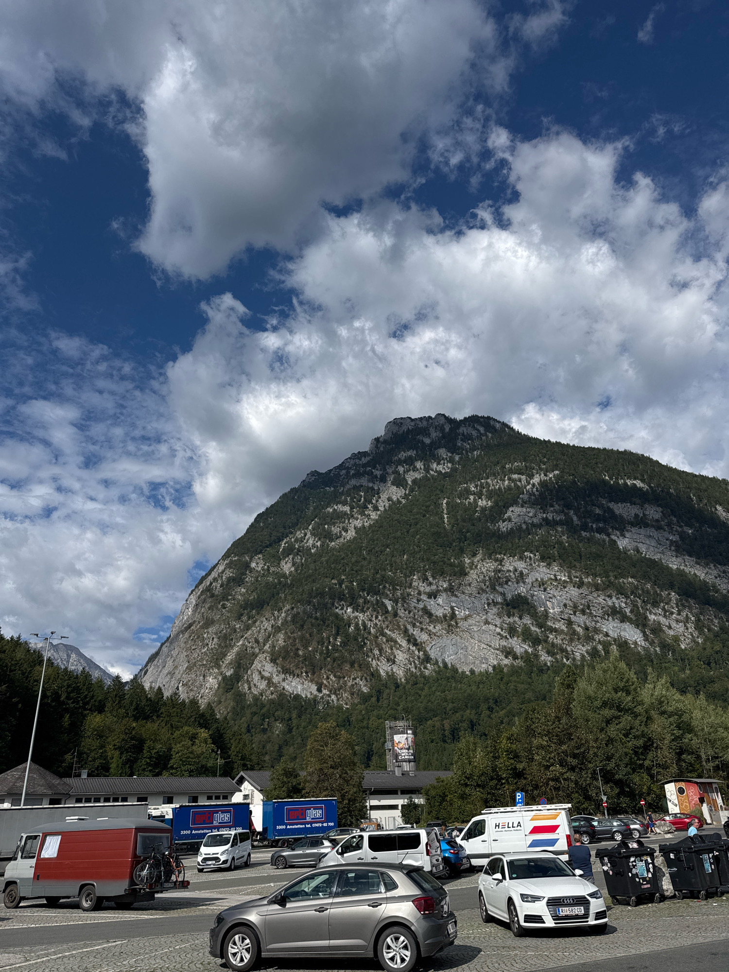 In the city of Torren, Austria, the scene is dominated by a large, rugged mountain that fills the background. The mountain's face is a mix of grey rock and dark green vegetation, with dramatic shadows cast across its surface by the clouds. In the foreground, a parking lot is filled with various vehicles. A gray hatchback is parked on the left, and to its right, a white sedan with an Austrian license plate is in motion, turning or parking. Further back, several trucks with blue and white livery display the text "mpt.glas" and phone numbers. A gray van with a red stripe is parked to the left, carrying bicycles on its rear. A white van with the HELLA logo is also visible. The environment suggests a busy area, possibly a rest stop or commercial hub, with a clear, partly cloudy sky overhead. The lighting indicates it is daytime.