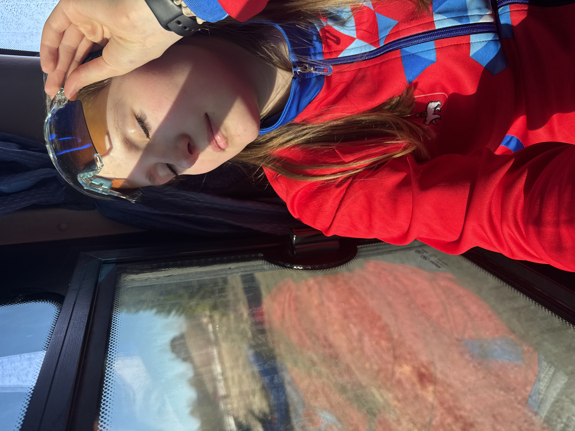 A person is lying down on a surface and holding up sunglasses. The person is wearing a red and blue jacket and has a black watch on their wrist. The surface is a vehicle windshield, and the reflection shows an unclear image of buildings. The location is Челва, Russia.