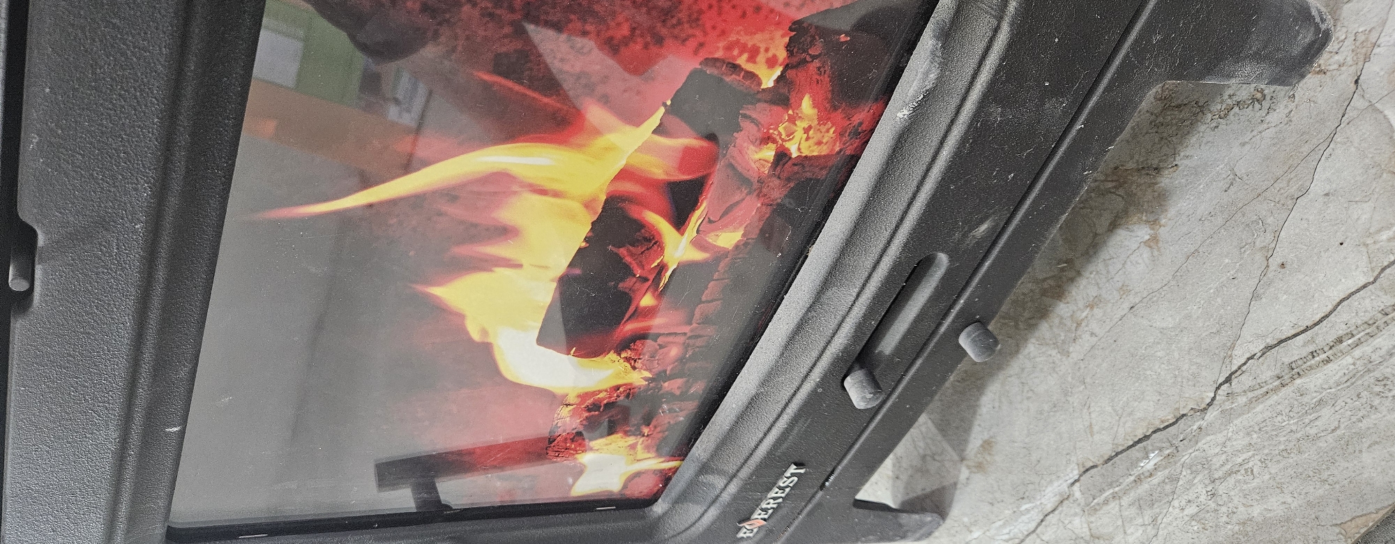 A close-up, vertical shot captures the glass door of a black, modern wood-burning stove. The stove has a textured, matte finish and two small, dark, knob-like controls are visible on the front panel. The word "EVEREST" is emblazoned in white on the lower left side of the stove. Behind the glass, a vibrant fire burns, with logs crackling amidst bright yellow and orange flames. The fire appears to be a simulated or decorative element, rather than a real fire, due to its static and uniform appearance. The stove sits on a mottled gray and beige floor that resembles marble or polished concrete, with visible cracks and veins running through it. The background is out of focus but suggests an indoor setting, possibly a room in Semey, Kazakhstan, where such stoves are common for heating purposes. There are no people present in the image.