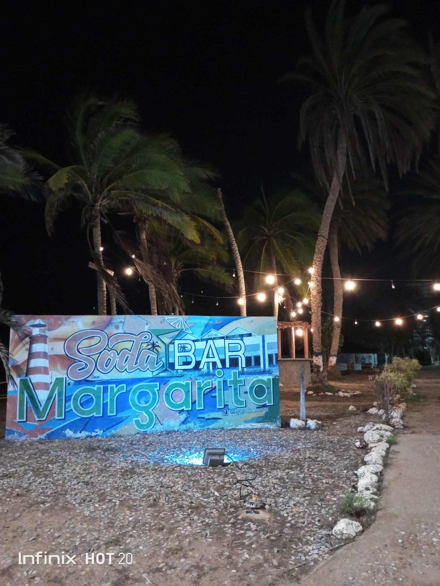 This nighttime photograph captures an outdoor bar or recreational area in Porlamar, Venezuela, under a dark, clear sky. The scene is brightly lit by numerous strands of warm incandescent string lights, draped between several tall palm trees that frame the setting and extend into the background.
The central subject is a large, colorful sign on the ground level, featuring stylized text. It prominently displays "Soda BAR" at the top, with "Margarita" in larger, vibrant letters below. In the lower right corner of the sign, smaller text appears to read "f sodabarmgta," likely a social media handle. A blue ground light sits in front of the sign, casting an upward glow onto it and the surrounding gravelly ground.
The ground consists of dirt and small stones, with a defined pathway on the right bordered by a line of white rocks. Sparse green vegetation grows alongside this border. In the mid-ground, a small wooden structure, possibly a bar or kiosk, is visible behind the sign and between the palm trees, emitting a subtle internal light. No people are visible in the image, but the arrangement of lights, the bar sign, and the general ambiance suggest a relaxed and inviting setting for evening leisure or social gatherings. A "Infinix HOT 20" watermark is present in the bottom left corner.
