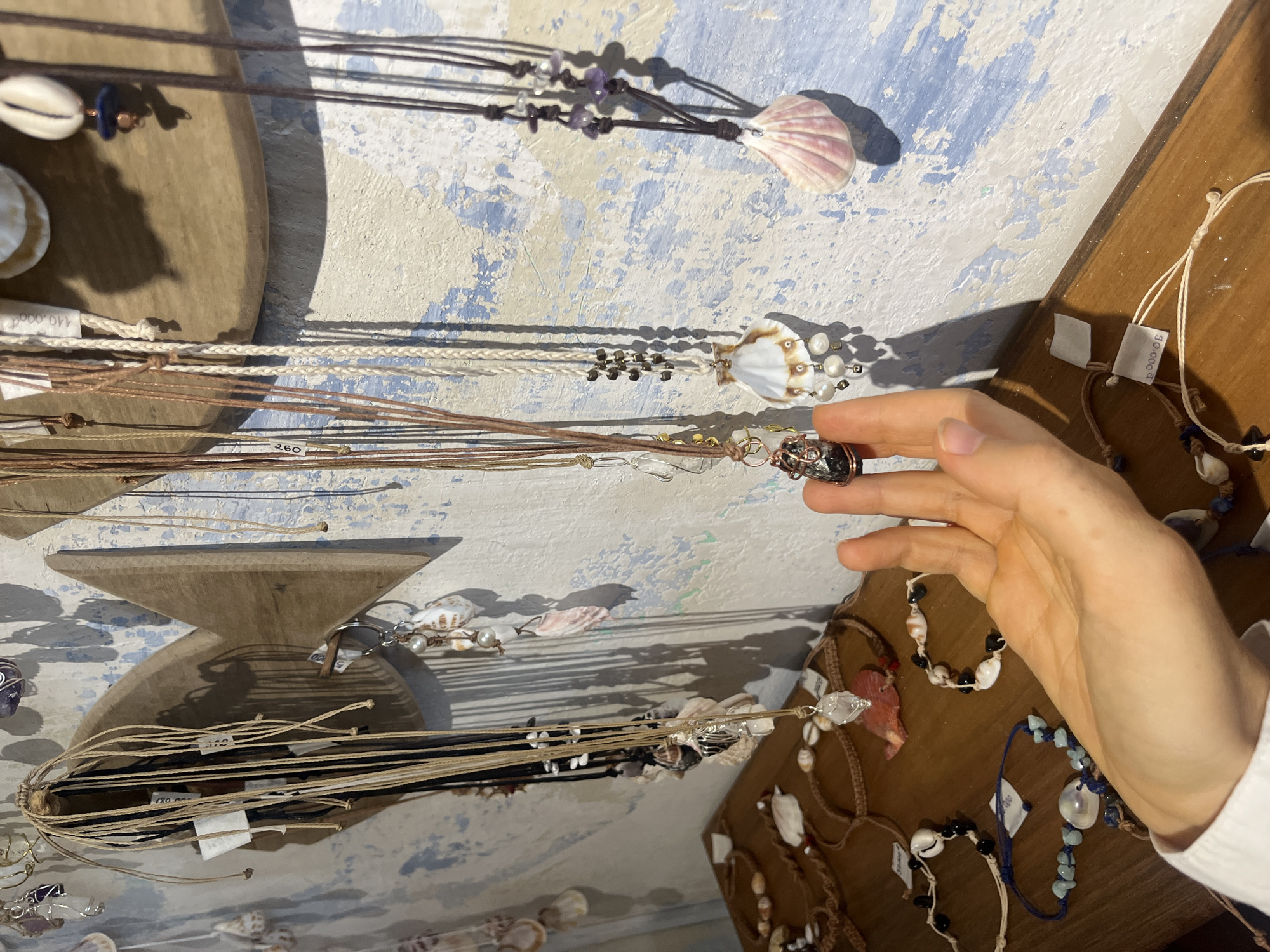 A person's left hand is in the foreground of the image, reaching towards a display of necklaces and bracelets. The jewelry is made of shells, pearls, beads, and crystals, and is suspended from a wooden fish-shaped display mounted on a textured blue and white wall. Several of the necklaces have price tags attached. Below the wall display, on a wooden shelf, are more bracelets and necklaces, some of which the hand is near. The scene appears to be in a shop. The location is identified as Phu Thuy Ward, Vietnam.