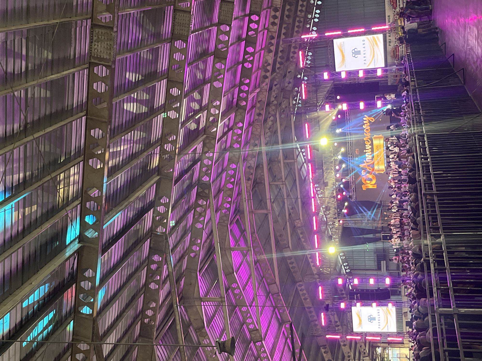 The image depicts an indoor event, likely a celebration or concert, held within a large, industrial-style venue with a corrugated metal roof supported by exposed metal beams and trusses. The space is illuminated by a combination of stage lighting, featuring vibrant pink and blue hues that cast colorful reflections on the ceiling. In the foreground, a crowd of people, mostly obscured by a metal railing, are gathered, facing a stage set up at the far end of the venue. The stage is adorned with various lighting rigs, large screens, and a banner that reads "10th Anniversary". There is a prominent screen on the left displaying the "PARKLAND" logo. A group of people, possibly performers or attendees, are visible on the stage. The overall atmosphere suggests a lively and celebratory event. The specific location in Pelang, Indonesia is not visually identifiable from the image.