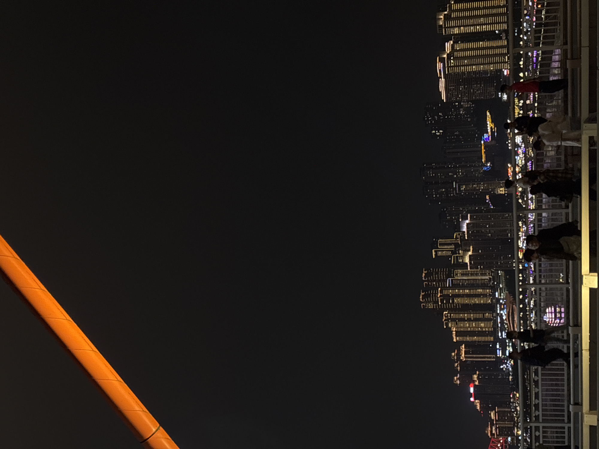 The scene depicts a nighttime view from a high vantage point in the Yuzhong District, China. The city skyline stretches out into the distance, illuminated by numerous lights from buildings, creating a vibrant urban landscape. People are present at the viewing point, observing the city lights, their forms are silhouetted against the night sky. In the foreground, a segment of an orange-colored structure is visible. The overall environment is dark, with the city lights providing the primary source of illumination.