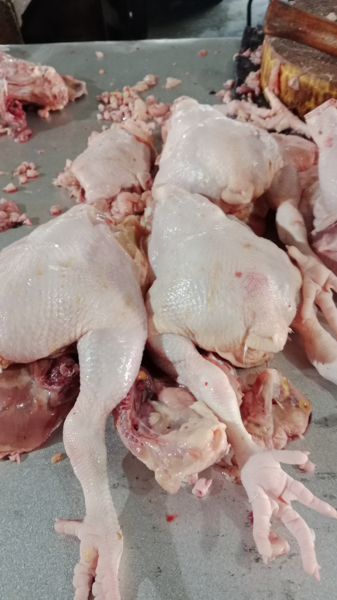 The image shows several whole, raw chickens that have been prepared for sale, likely in a market in Denpasar, Indonesia. The chickens are laid out on a grey, mottled surface, possibly a countertop or a table. Some of the chickens are intact, while others appear to have been partially butchered, with exposed meat and bones. Various pieces of raw chicken, including wings, legs, and potentially other internal organs, are scattered around the whole birds. The chickens are pale pinkish-white, with some showing slight yellowing of the skin. The scene suggests a commercial setting where poultry is processed and sold. The presence of a large wooden cleaver and cutting block in the upper right corner further supports the interpretation of this being a food preparation or market environment. There are no people visible in the image.