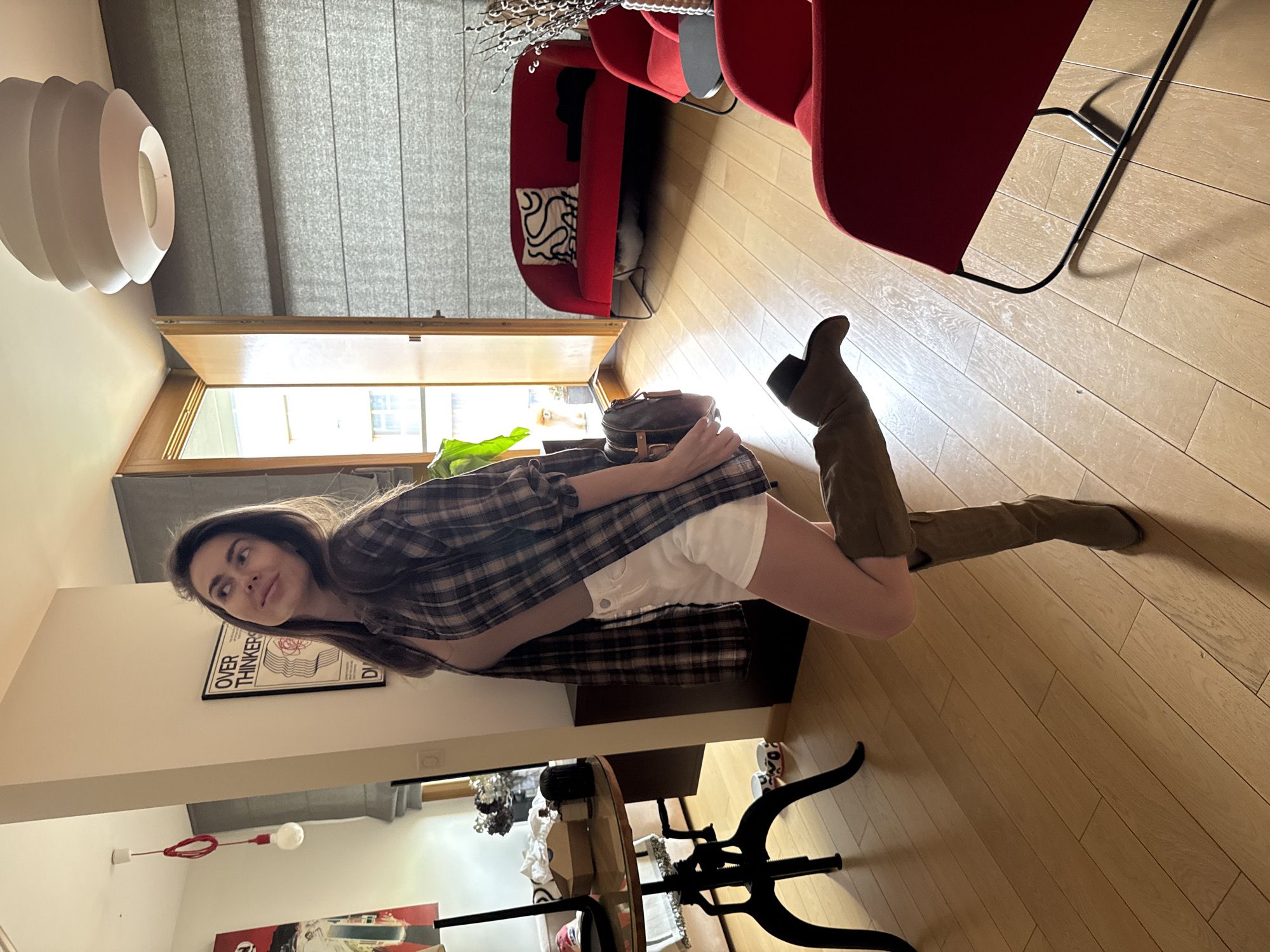 A woman is posing in a room. The room has light wood floors, a red chair and sofa, and a table. The woman is wearing a plaid shirt, white shorts, and tall brown boots. She is facing left, and her leg is extended in front of her. There is a light fixture hanging from the ceiling and a poster on the wall. The location is Unknown, Unknown.