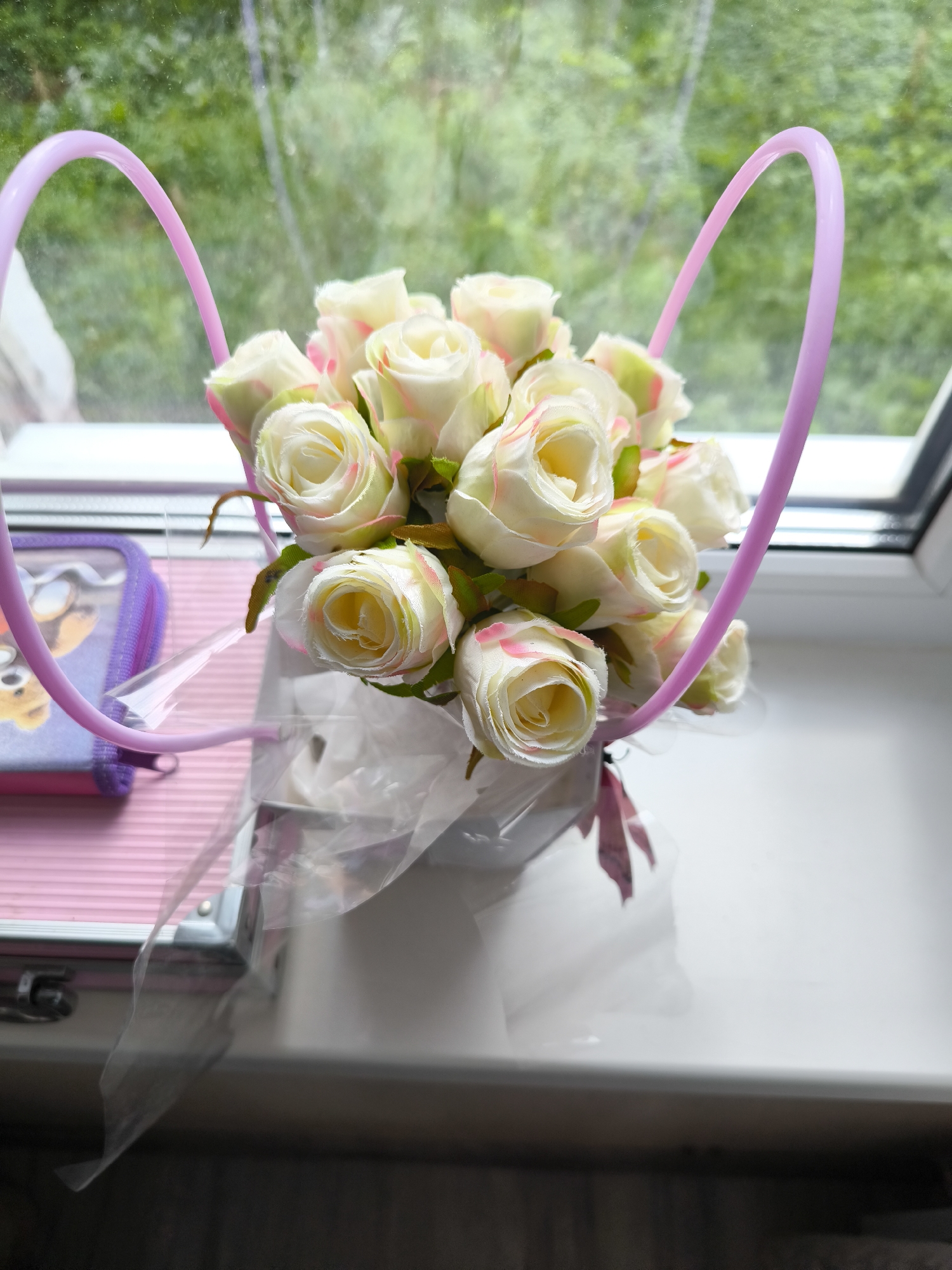 A bouquet of artificial white roses with pink tips is displayed in a white container with a pink ribbon. The container is placed on a white windowsill, with a window behind it looking out onto greenery. To the left of the roses is a small pink zippered pouch with a cartoon character on it. The scene appears to be indoors, possibly in a home or room, with natural light coming from the window. The overall impression is one of simple decoration or a gift arrangement.