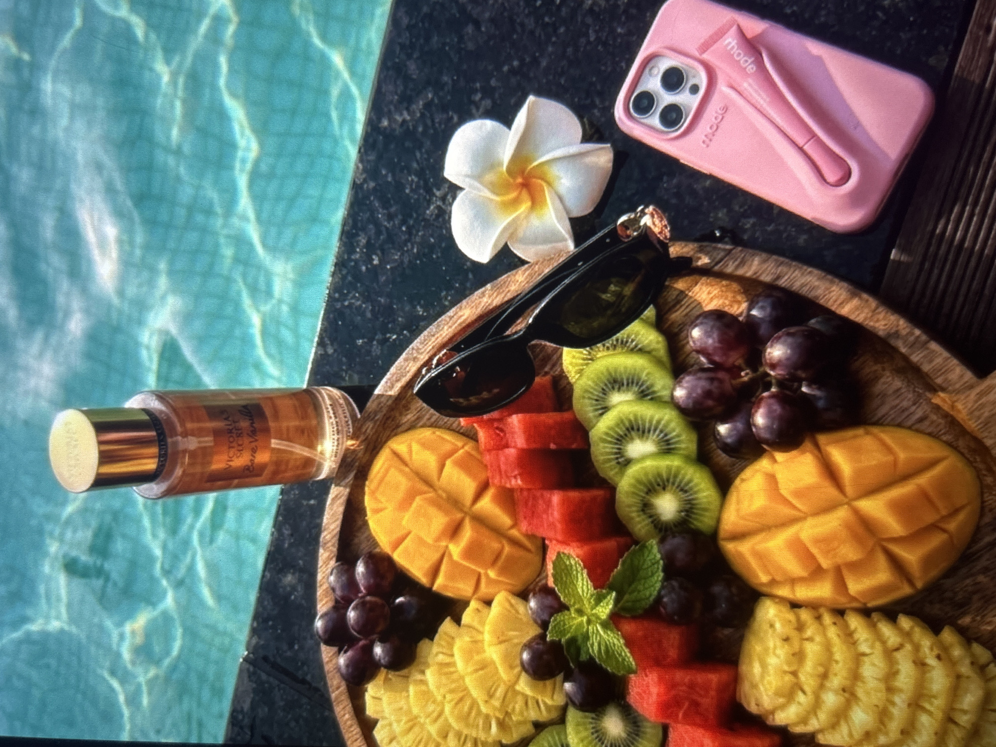 The image shows a fruit platter with pineapple, mango, watermelon, kiwi, and grapes arranged on a wooden board. The platter is placed on a dark surface next to a swimming pool. A bottle of Victoria's Secret Bare Vanilla body mist, a pair of sunglasses, a white flower, and a pink phone case with a lip balm are also visible. The scene appears to be set outdoors, with the water of the pool in the background reflecting sunlight. There are no people or animals present in the image. The location in Knezha, Bulgaria is not identifiable from the image.