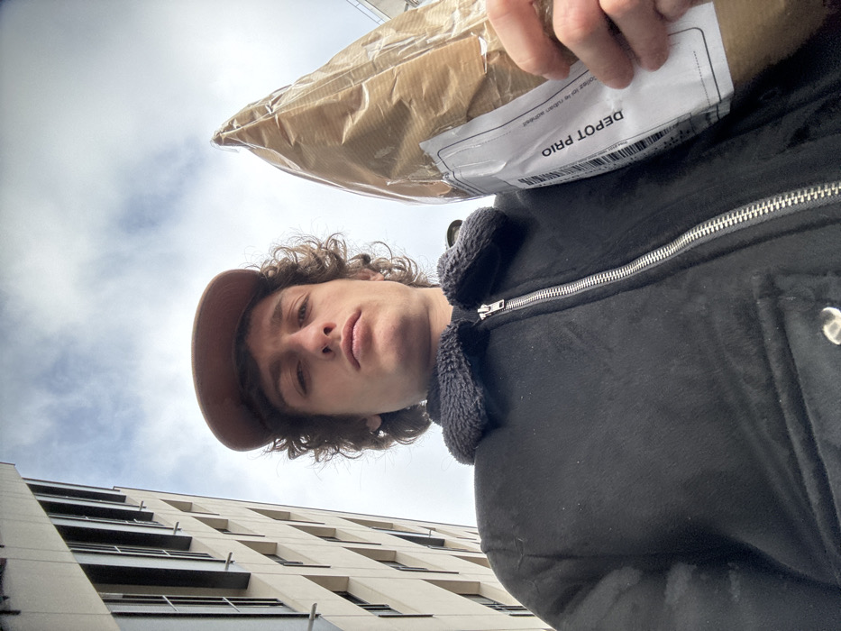 A low-angle selfie captured in Joinville-le-Pont, France, depicts a male individual from the chest up, positioned against a backdrop of urban architecture and sky. The individual has medium-length curly brown hair and wears a light brown baseball cap. He is dressed in a black jacket featuring a dark, possibly fleece, collar and a prominent silver zipper running down the front. He looks directly at the camera with a neutral expression. In his left hand, on the right side of the frame, he holds a package wrapped in light brown paper and clear plastic. A white shipping label, vertically oriented, is affixed to the package, displaying a barcode and the text "DEPOT PRIO." The background on the left features a portion of a tall, modern building with light-colored facade panels and multiple rectangular windows. Above the building and the subject, a partly cloudy sky is visible.
