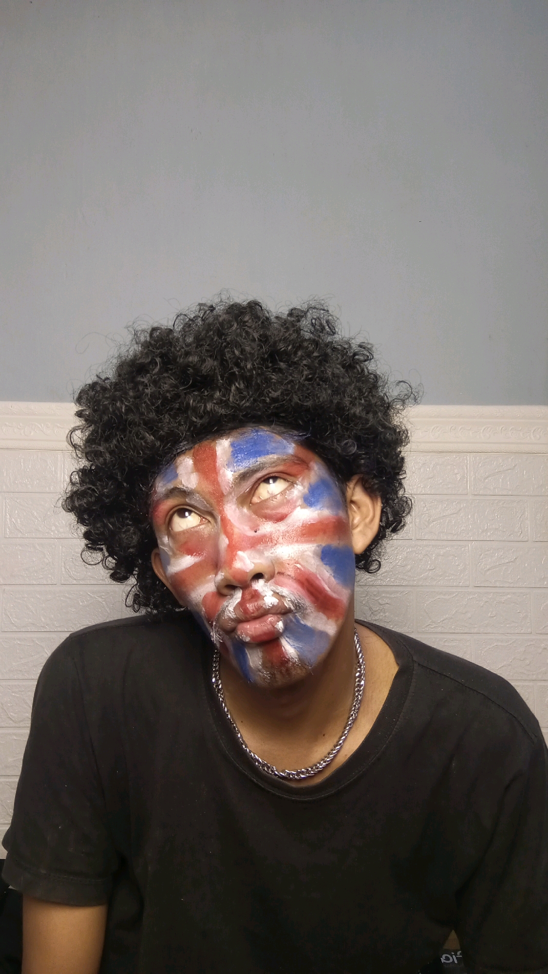 A person with dark, curly hair and face paint resembling the Union Jack flag poses for a photo. The person is wearing a black t-shirt and a silver chain necklace. They are looking up and to the left, with their mouth slightly parted. The background appears to be a light blue wall with a white brick pattern. The image is a selfie taken from a medium shot, focusing on the person's face and upper body. The location is identified as Kebumen, Indonesia.