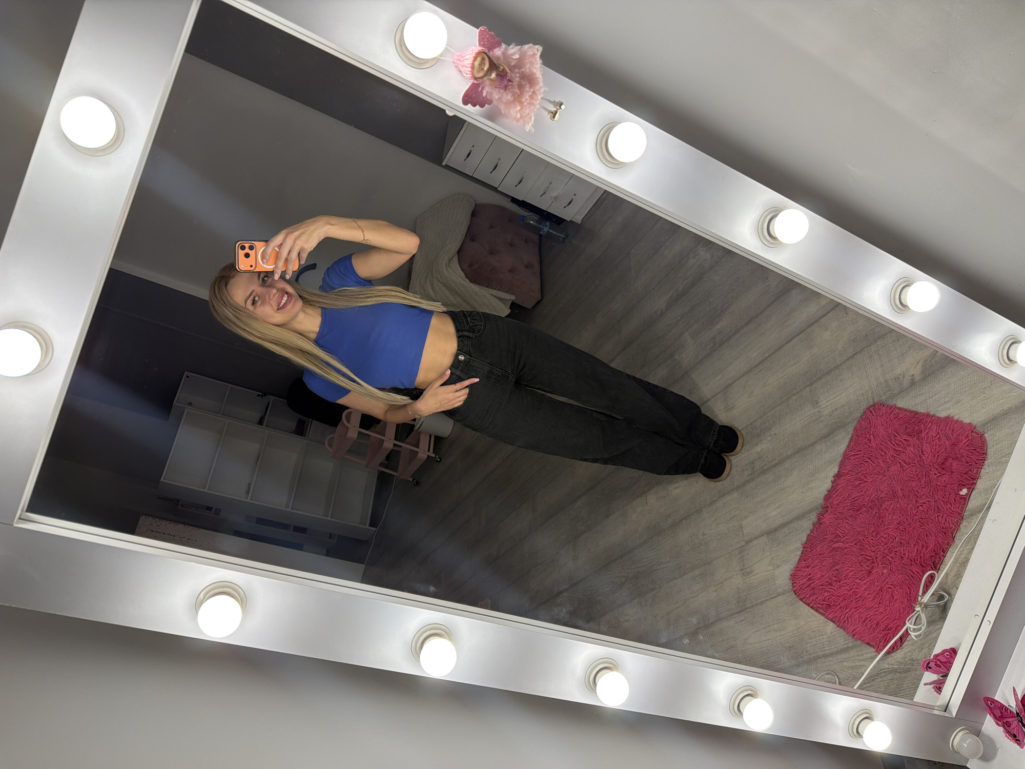 A young woman with blonde hair is taking a mirror selfie in what appears to be a room with a vanity mirror. She is wearing a blue crop top and black jeans. The mirror is illuminated by several light bulbs. In the reflection, a pink fuzzy rug is visible on the floor, as well as a pink trolley cart, a chest of drawers, and a tufted pink ottoman. A small fairy figurine is attached to the side of the mirror. The room has a gray wall and a wooden floor. The image does not contain any information that would allow for the determination of its location in Kyiv, Ukraine.