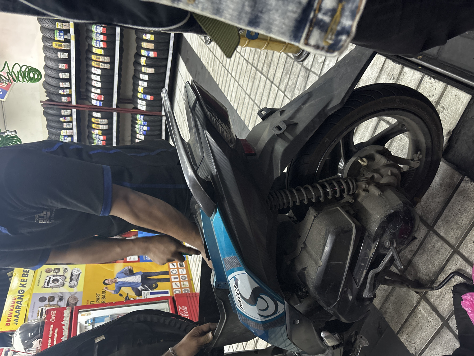 In North Jakarta, Indonesia, a mechanic is working on the engine cover of a teal and black scooter. The scooter is parked on a checkered tile floor. In the background, shelves are filled with tires. A red Coca-Cola cooler is visible to the left. The mechanic wears a dark blue shirt with a blue collar and black pants. The scooter has a "Mio" logo on its side.