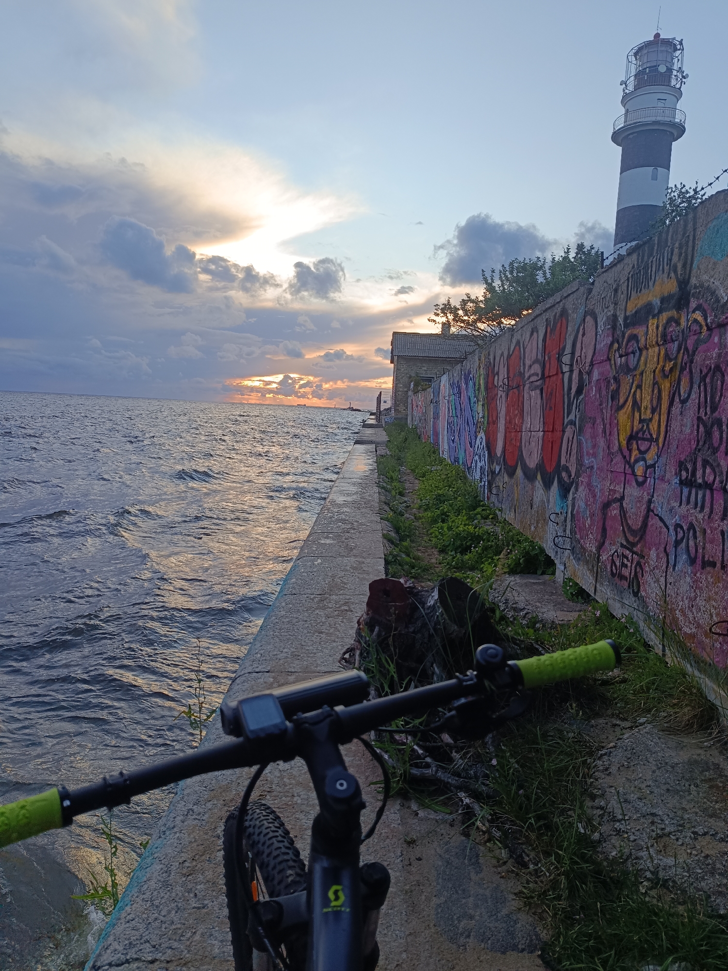 The image shows a view of a lighthouse and the sea at sunset, with a bicycle in the foreground. The lighthouse is tall and striped with black and white. It is situated on top of a concrete wall that is covered in colorful graffiti. The sea is choppy and reflects the warm colors of the sunset. The sky is cloudy with patches of orange and yellow from the setting sun. The bicycle's handlebars and front wheel are visible, indicating it is parked at the scene. The overall impression is of a picturesque, yet somewhat gritty, urban waterfront setting.