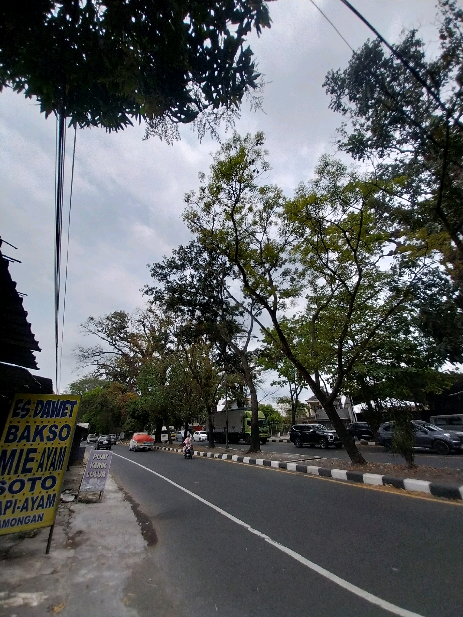 A street scene is captured in Tamanmartani, Indonesia, under an overcast sky. On the left side of the image, a weathered yellow sign displays food items in black lettering, including "ES. DAWET", "BAKSO", "MIE AYAM", "SOTO", and "API-AYAM LAMONGAN". Adjacent to the sign, a smaller sign reads "PIJAT KERIK LULUR". A paved sidewalk runs alongside the road, with power lines overhead.
The road itself is asphalt, marked with a white center line and a black and white striped curb. Several vehicles are visible: a green truck, a red and white truck, a black SUV, a dark colored SUV, and a grey SUV. A person on a motorcycle is also present. Lush green trees line the right side of the road, their branches reaching towards the sky. Some buildings are visible in the background, peeking through the foliage. The overall atmosphere is that of a typical roadside scene in a town or city.