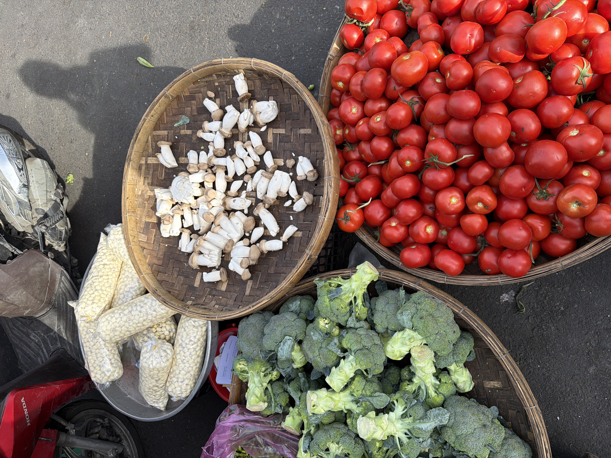 This is an outdoor, eye-level, medium shot of a produce stall in Nha Trang, Vietnam. The scene is dominated by four baskets of fresh vegetables and fruits. In the foreground, to the right, a woven basket overflows with ripe red tomatoes. To the left, another woven basket is filled with bright green broccoli florets. Above these, a round, woven tray holds a pile of sliced white mushrooms. Behind the mushrooms, a metal basin is filled with numerous small, white, bean-like items sealed in clear plastic bags. A red Honda motorcycle is partially visible on the left edge of the frame, and a person's shadow is cast on the asphalt ground to the right. The ground is dark asphalt, suggesting a street or market setting. The overall impression is one of a vibrant, open-air market.