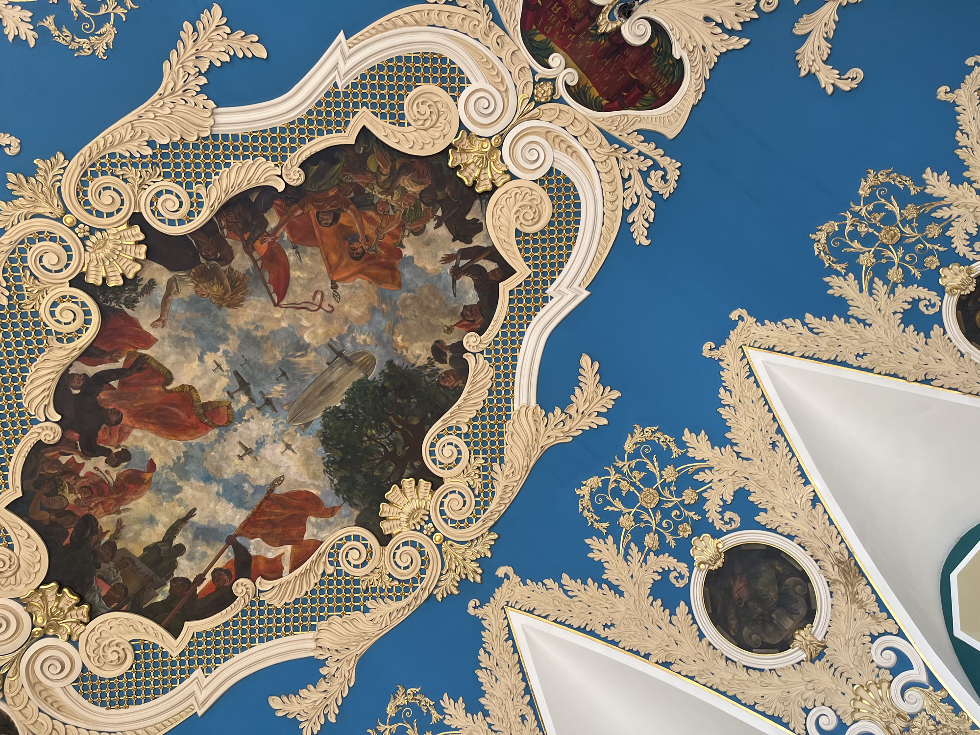 The image captures a portion of the ornate ceiling within the Mayakovskaya Metro Station in Moscow, Russia. The ceiling features a pale gold decorative frame surrounding a fresco. The fresco depicts a sky scene filled with numerous figures holding red flags. Overhead, multiple airplanes and a large dirigible are painted, suggesting an aerial procession. The scene is enclosed within a stylized white and gold border. The background is a vibrant blue color and features a gold floral pattern. A circular portrait can also be seen within the decorative frame. A white, angled, architectural feature is partially visible in the lower right. The overall style is that of Socialist Realism.
