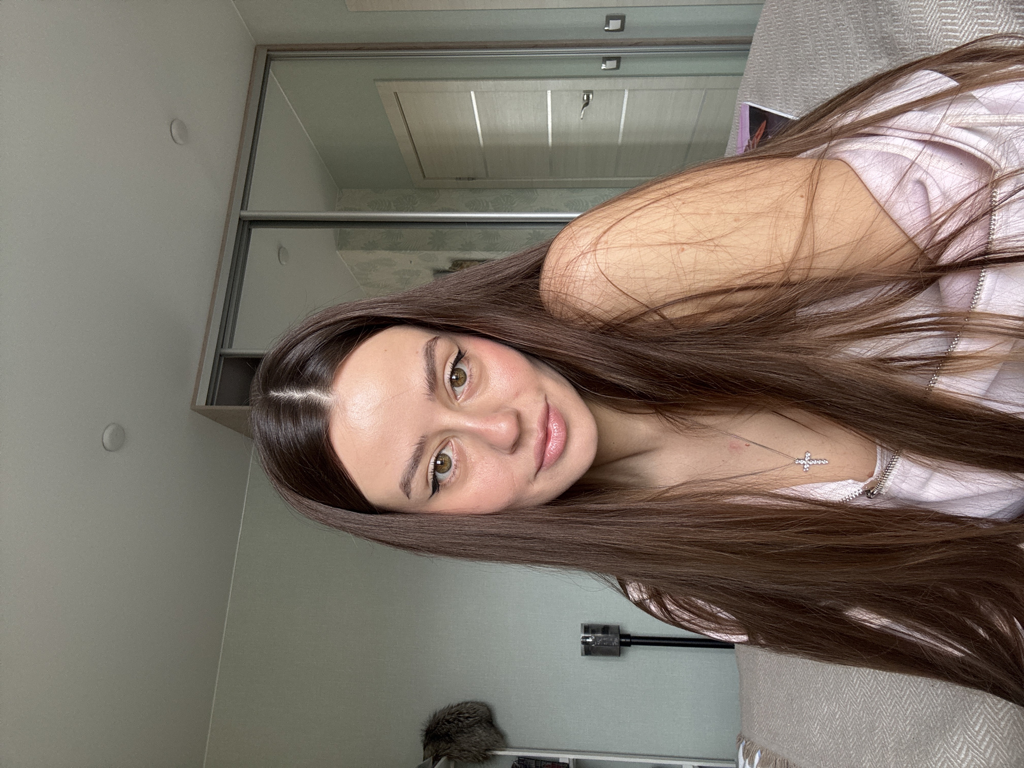 A young woman with long, brown hair and green eyes is looking at the camera with a slight smile. She is wearing a light-colored shirt with a silver chain and a cross necklace. The room appears to be a bedroom, with sliding closet doors and a bed visible in the background. A vacuum cleaner is affixed to the wall. The location is not indicated in the image.