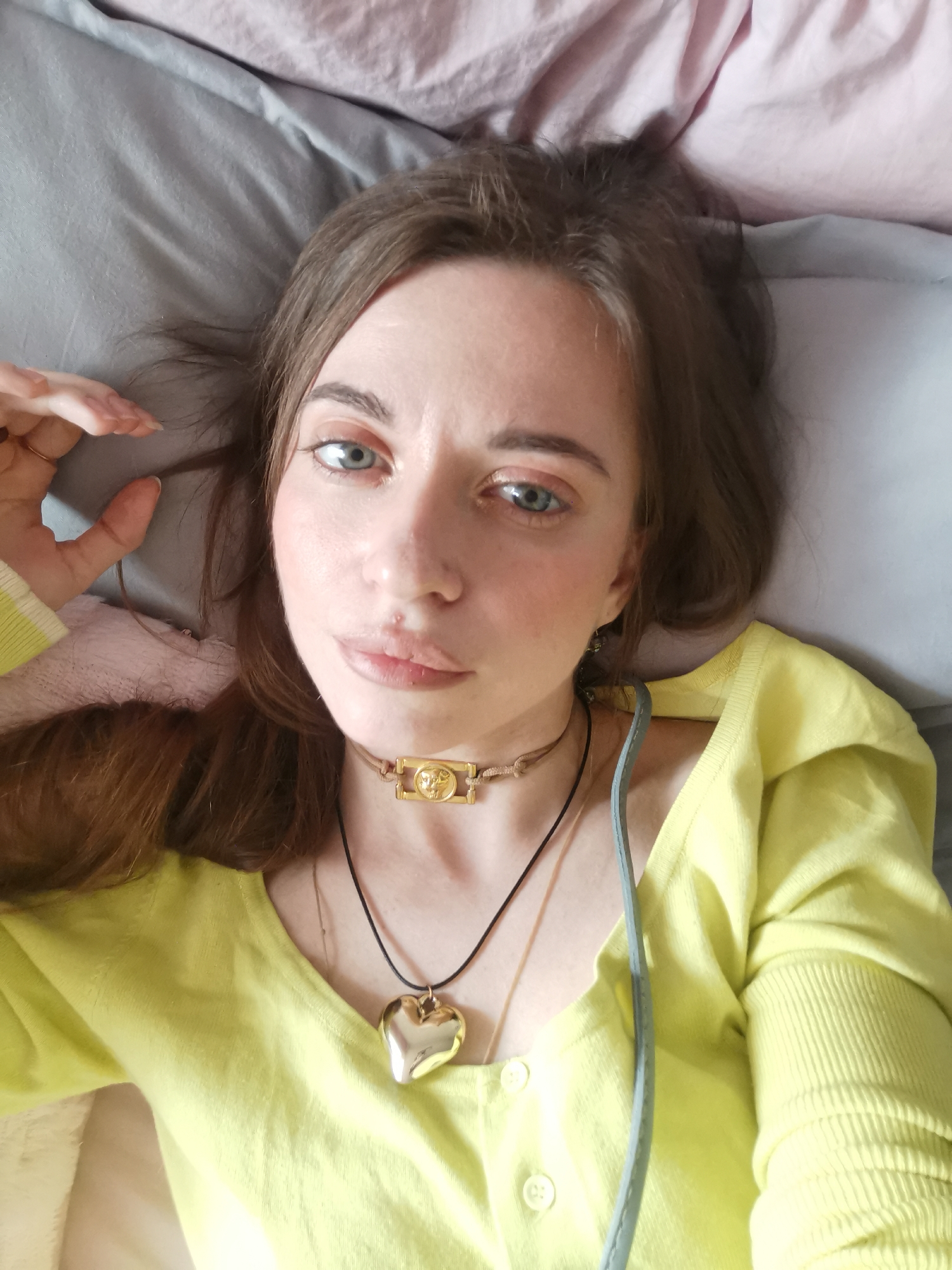 A young woman with light skin and blue eyes lies on a gray pillow, wearing a yellow cardigan, with a gold heart pendant necklace and a choker with a gold rectangular charm. Her brown hair is spread around her head, and she is looking directly at the camera. A pink blanket is partially visible in the background. The location is unknown.