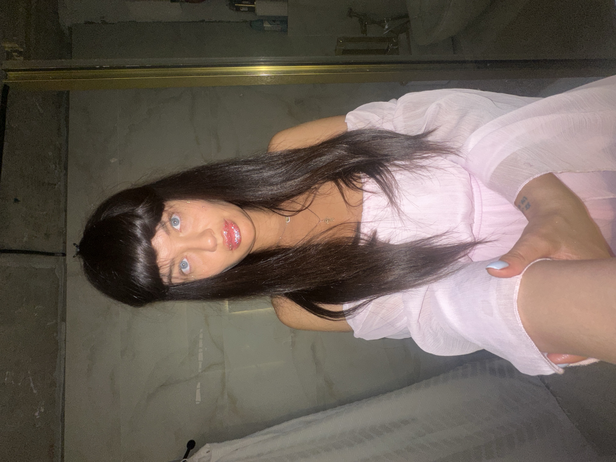 The image displays a head-and-shoulders portrait of a person, likely female, with long, straight, dark hair and bangs that frame their forehead. The person has striking blue eyes that gaze directly at the camera. Their lips are painted with a glossy, sparkly pink lip product. They are wearing a light pink garment, possibly a dress or a flowy top, made of a sheer or lightly textured fabric. A delicate necklace with a small pendant is visible around their neck. One hand is partially visible, showing light blue nail polish on the fingernails, holding up the fabric of their top/dress. A faint, unreadable tattoo or marking is present on the inner part of their forearm.
The setting appears to be an indoor space, likely a bathroom, characterized by textured, greyish-beige walls. A gold-colored metallic frame, possibly belonging to a shower door or a large mirror, runs horizontally across the upper portion of the background. Reflections of overhead lights and indistinct bathroom fixtures can be seen in the surface behind this frame. In the lower right corner, a white, textured towel hangs, suggesting a bathroom environment. The lighting is somewhat dim, indicating artificial illumination, possibly from overhead sources reflected in the background. The person's direct gaze suggests they are posing for a photograph or taking a selfie. No discernible text is visible other than the faint marking on the arm.